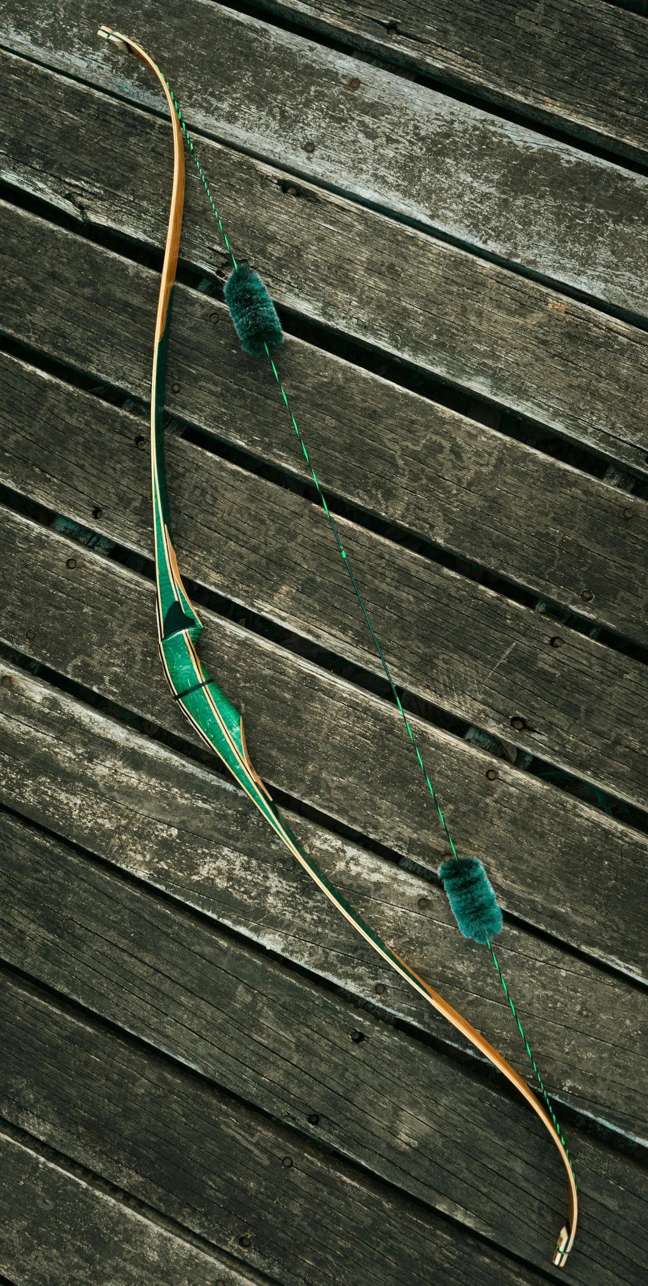 A traditional recurve bow with a green and wooden design, resting on weathered outdoor wooden planks. Attached to the bowstring are two teal fuzzy finger guards.