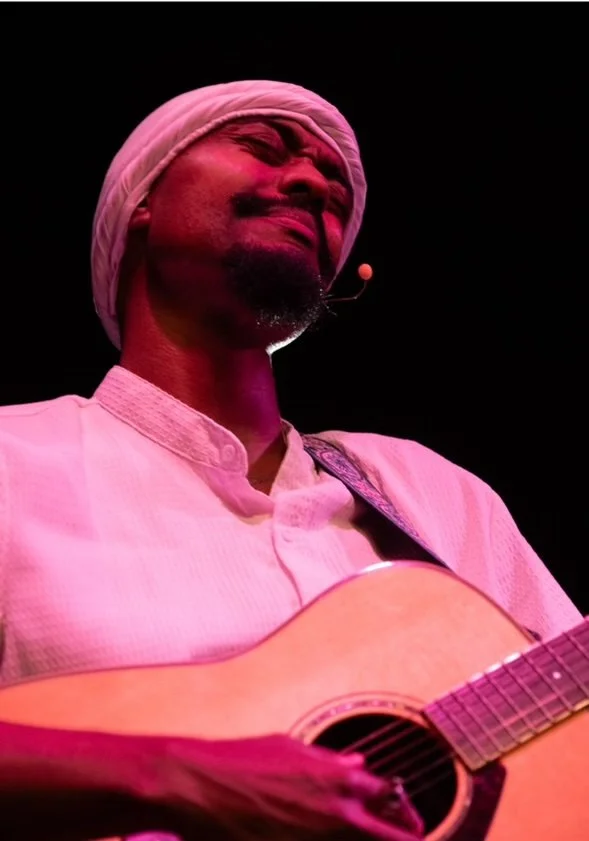 A man wearing a white turban and traditional white attire passionately plays an acoustic guitar on stage, illuminated by pink stage lighting.