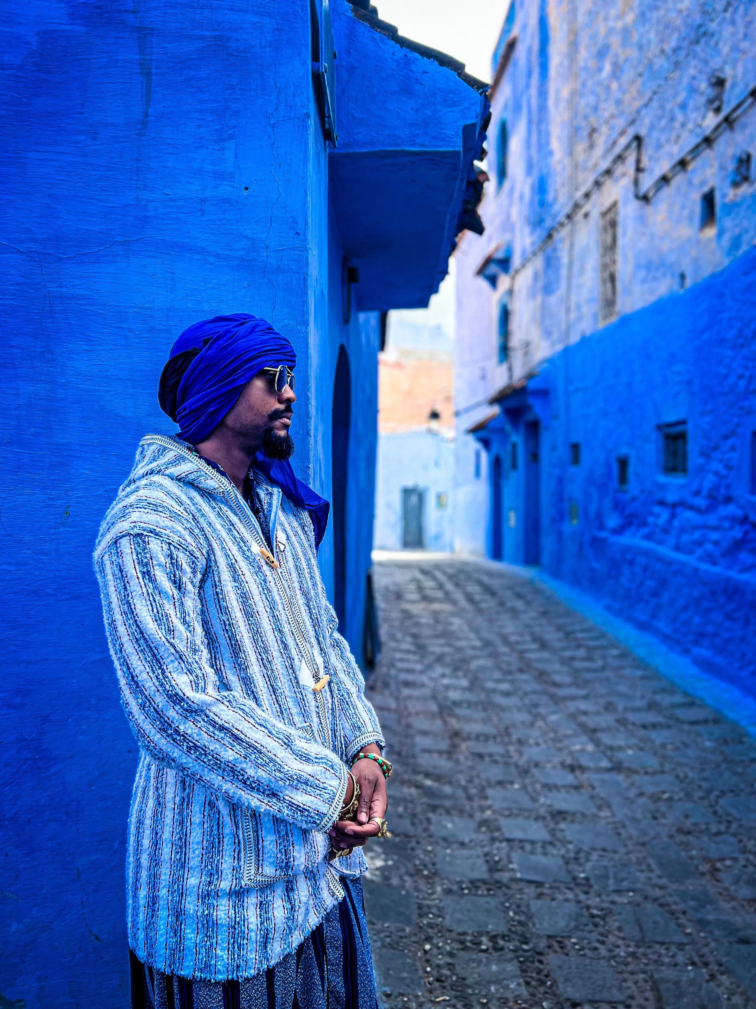 A man wearing a blue headwrap, sunglasses, and a patterned coat stands with his hands clasped in a narrow alleyway painted blue, with cobblestone pavement and blue walls on either side.