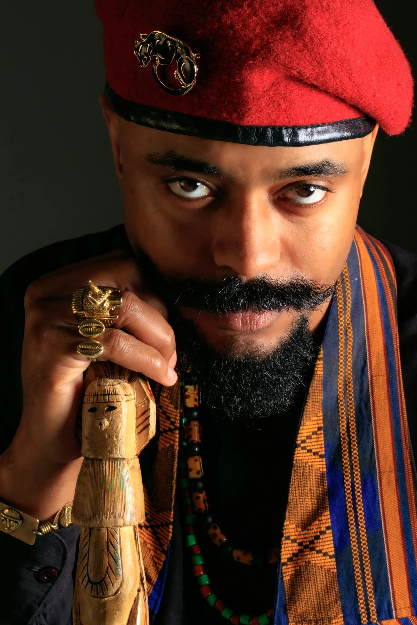 A man dressed in traditional African attire holding a carved wooden staff and wearing a red beret with a black panther emblem. He has a beard, mustache, and wears gold rings and jewelry.