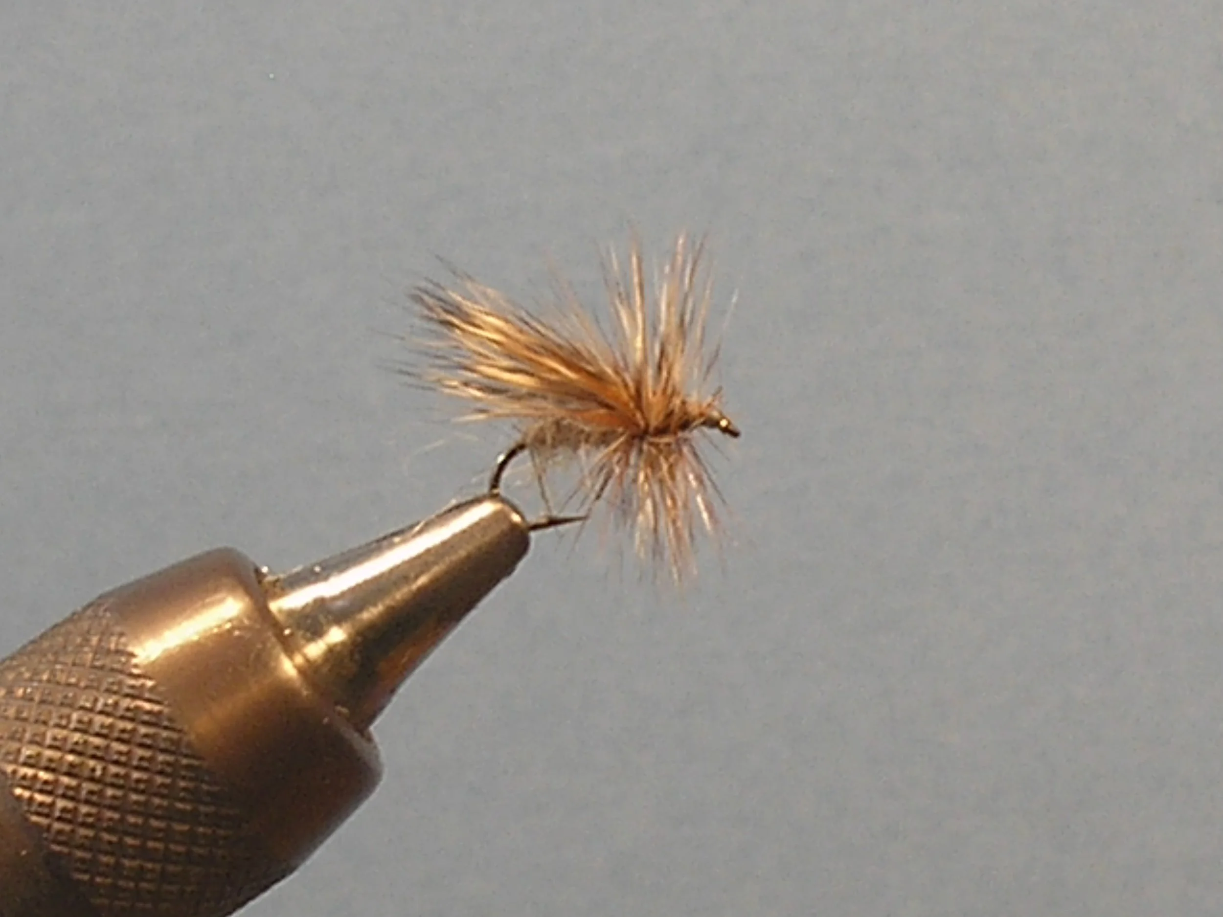 Close-up of a fishing fly with brown and tan fur, held in a metal vise against a plain gray background.