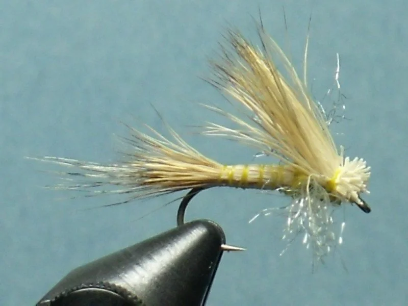 Close-up of a handmade fishing fly with beige feathers and a black hook on a light blue background.