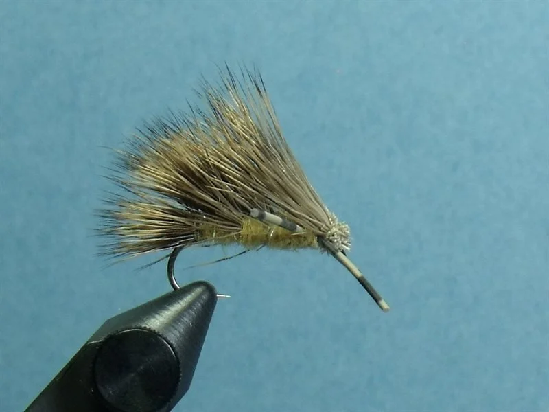 Close-up of a fishing fly designed to resemble a bait fish or insect, mounted on a hook and held in a display tool against a plain blue background.