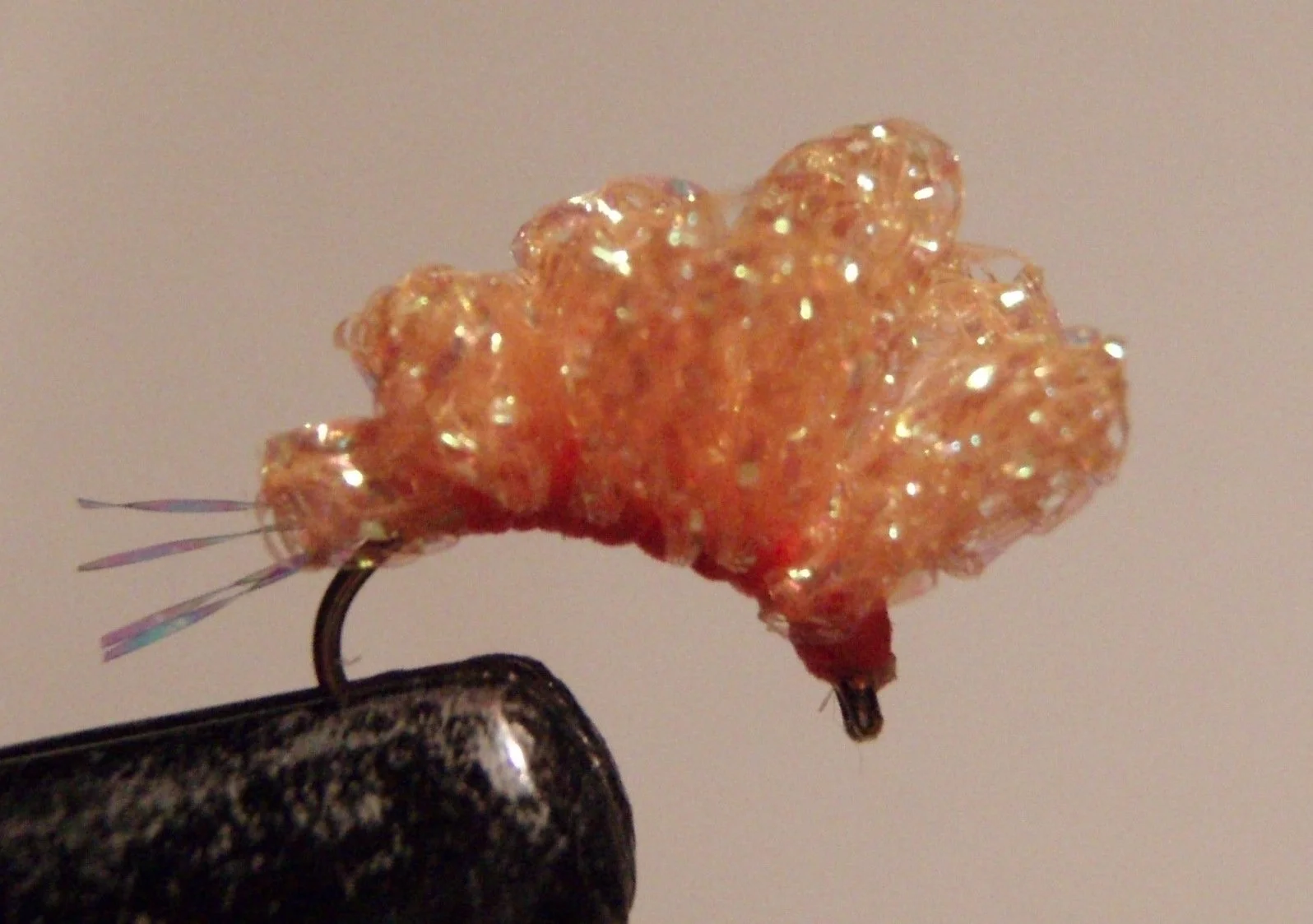 Close-up of a fishing fly, an artificial lure with orange, glittery, foam-like body, mounted on a barbed hook, used for fly fishing.