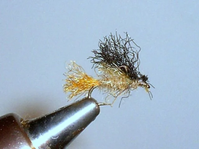 Close-up of a fishing fly or lure with black and yellow fibers, mounted on a metal hook against a light background.