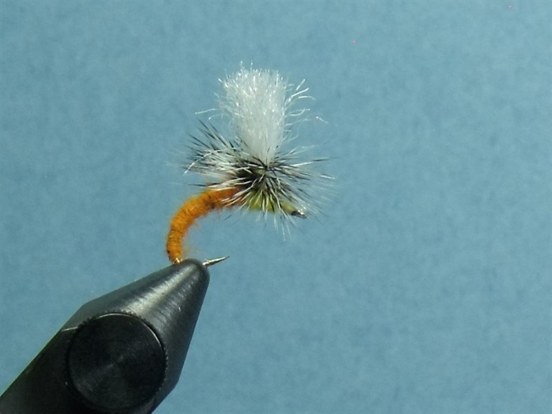 Close-up of a fishing fly lure with a white and black feathered head, attached to a hook on a black vise against a blue background.