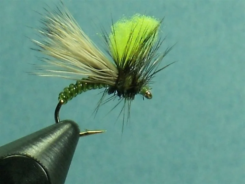 Close-up of a fishing fly with a black hook, green body, and fluffy yellow and black tail, held in a black vice against a light blue background.