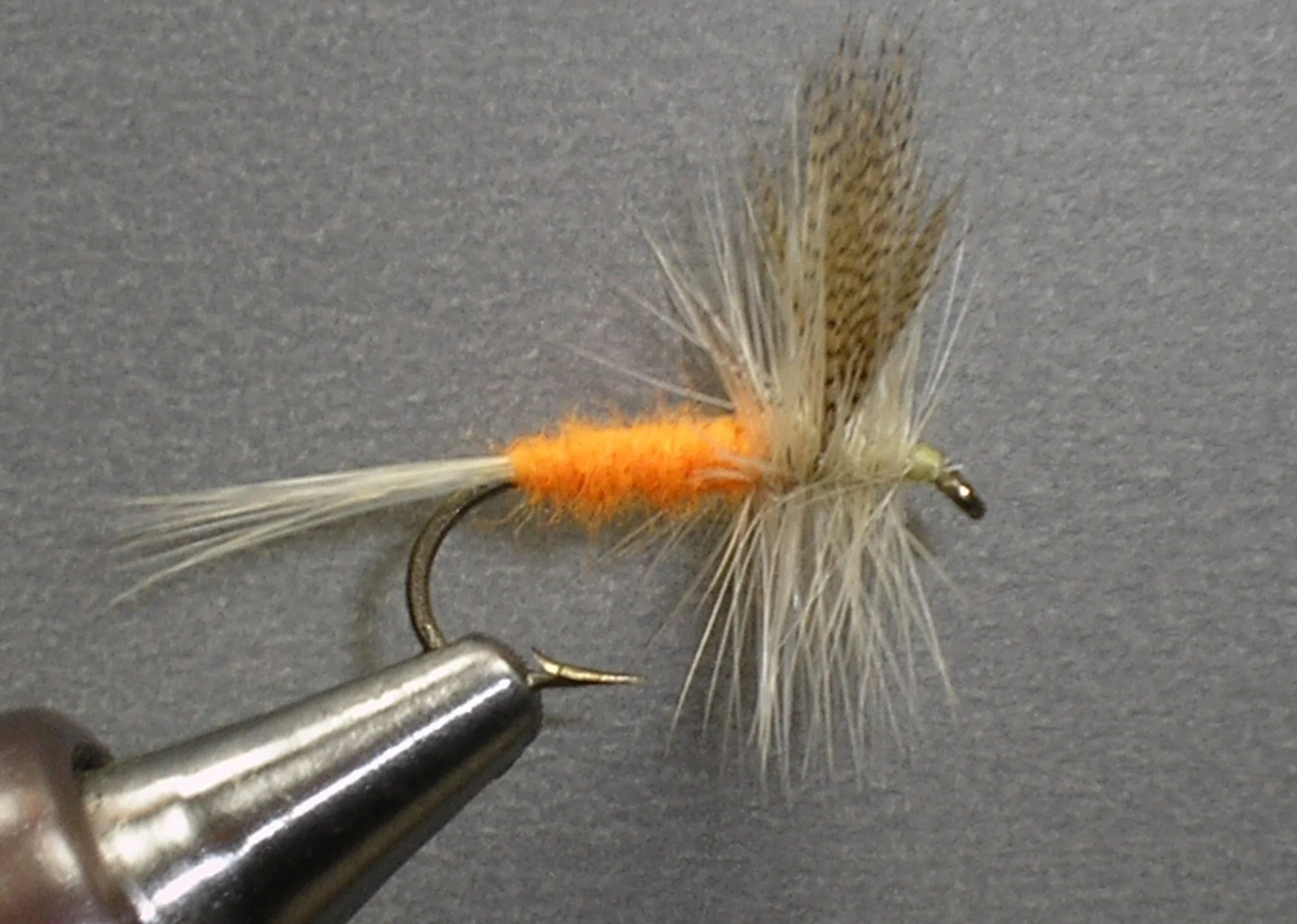 Close-up of a fishing fly lure with orange fuzzy body, beige and brown feathers, and a curved metal hook, held in a metallic holder on a gray background.