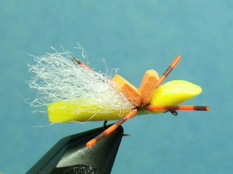 Close-up view of a fishing fly with yellow, orange, and white materials, held by a black tool against a blue background.