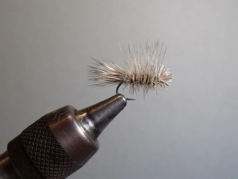 Close-up of an artificial fishing fly tied to a metal hook, held by a vice or tool.