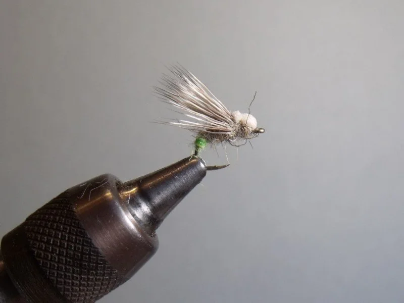 Close-up of a fishing fly with a gray body, white feathered wings, and a small green feature, mounted on a metal hook held in a vise against a plain background.
