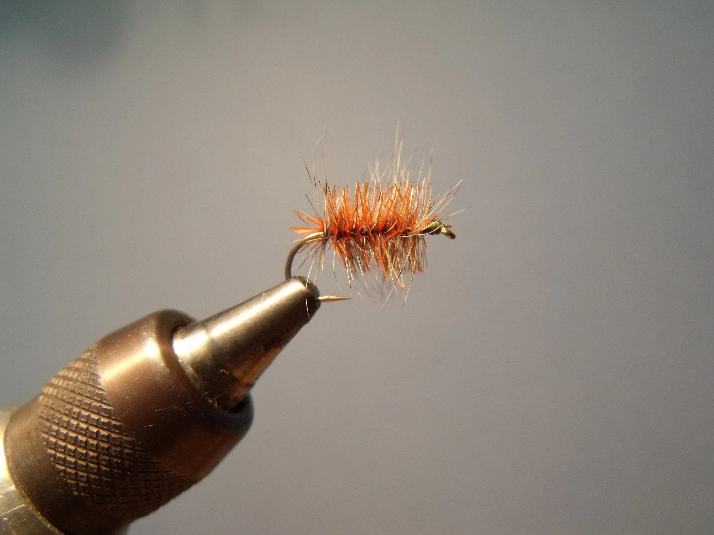 Close-up image of a handmade fishing fly with orange and brown feathers, mounted on a metal hook, being held by a needle-nose plier against a blurred gray background.