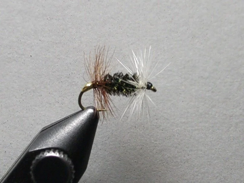 Close-up view of a fishing fly fishing lure with brown, black, and white fibers, mounted on a metal hook, against a gray background.