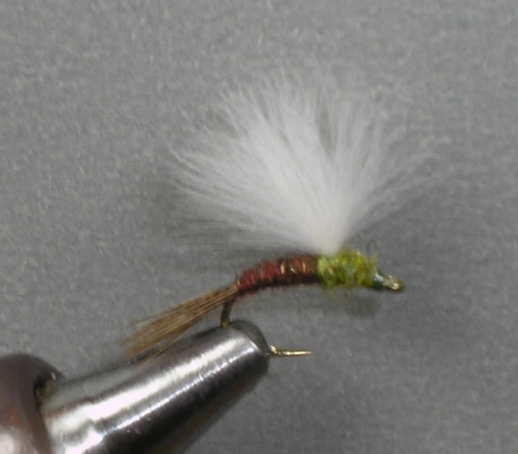 Close-up of a fishing fly with a white feather, brown tail, green body, and hooked onto a metallic tool on a grey background.