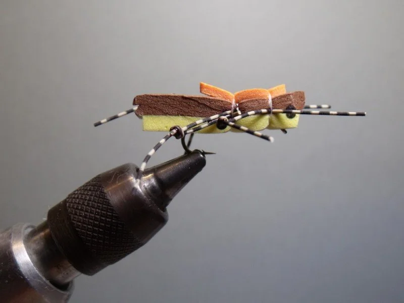 Close-up of a handmade insect craft resembling a fly, made from foam, wire, and wood, held with a tool against a gray background.