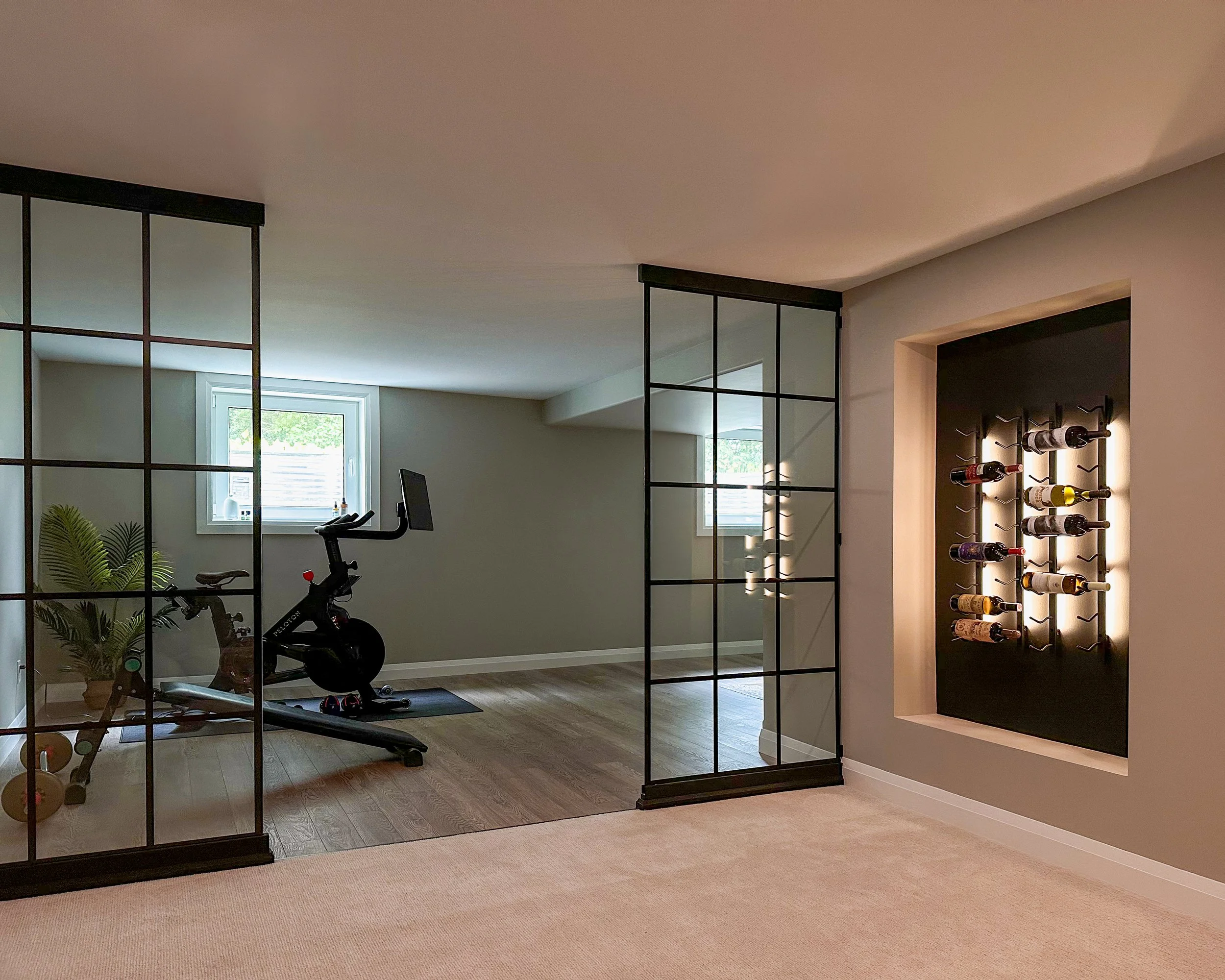 Basement renovation featuring a home gym behind glass partition walls and a built-in backlit wine rack, illustrating lifestyle-focused design.