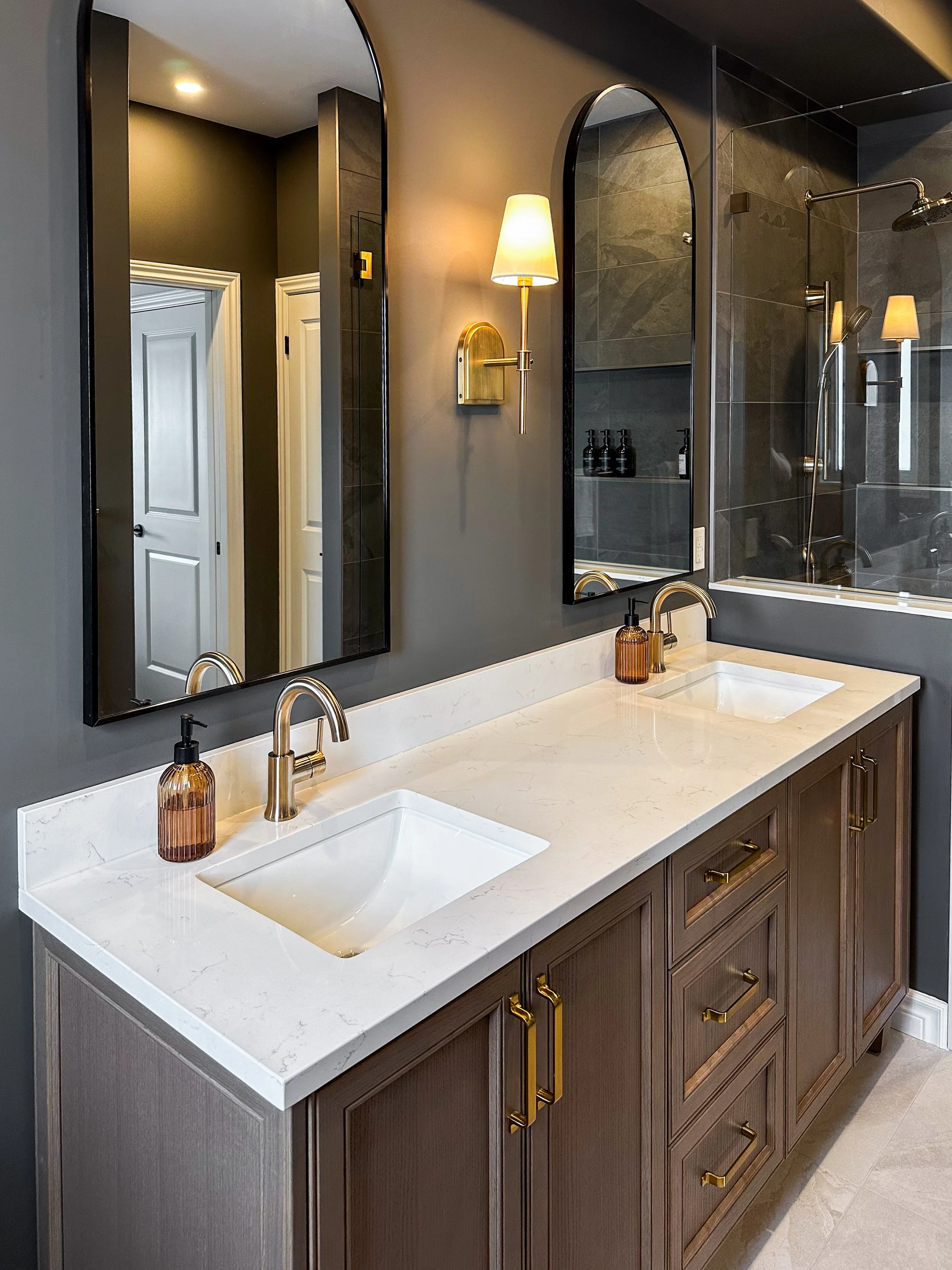 A full view of the dual bathroom vanity setup showcasing perfect symmetry. The warm wood cabinets, matching black arched mirrors, and brass wall sconces create a stunning organic modern aesthetic.