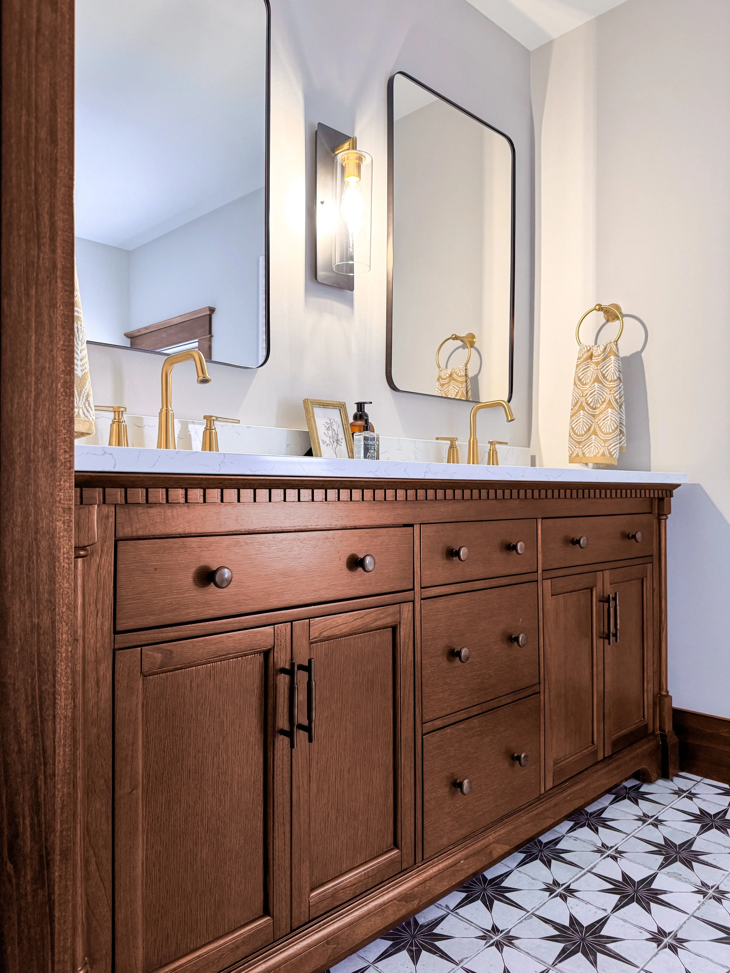A luxurious custom walnut double bathroom vanity with brass faucets, and a black and white starburst patterned tile floor in a newly renovated bathroom.