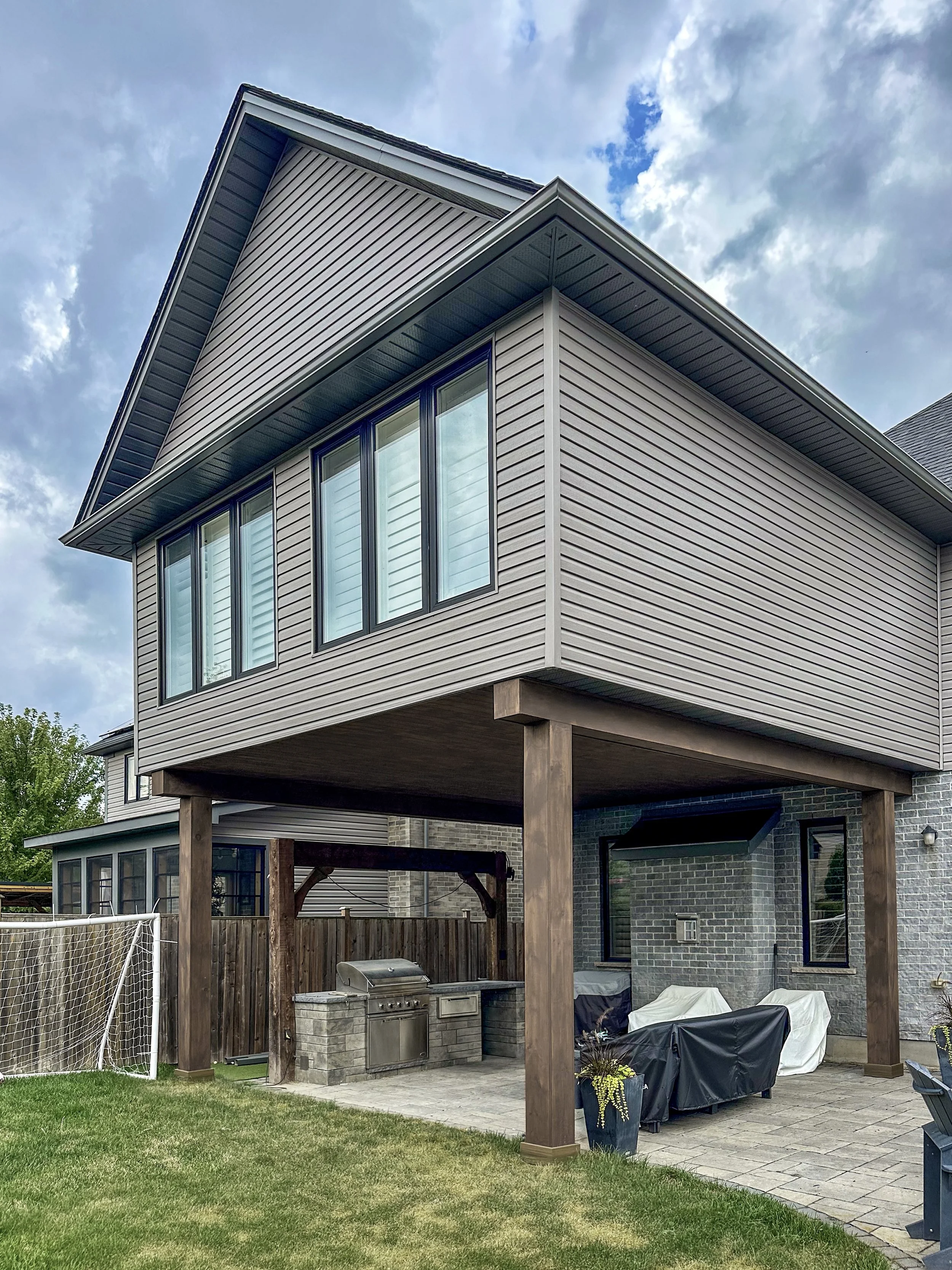 The completed second-story addition features colour matched siding and large windows, while the beautifully wood-wrapped structural columns transform the patio below into a fully covered outdoor living space.