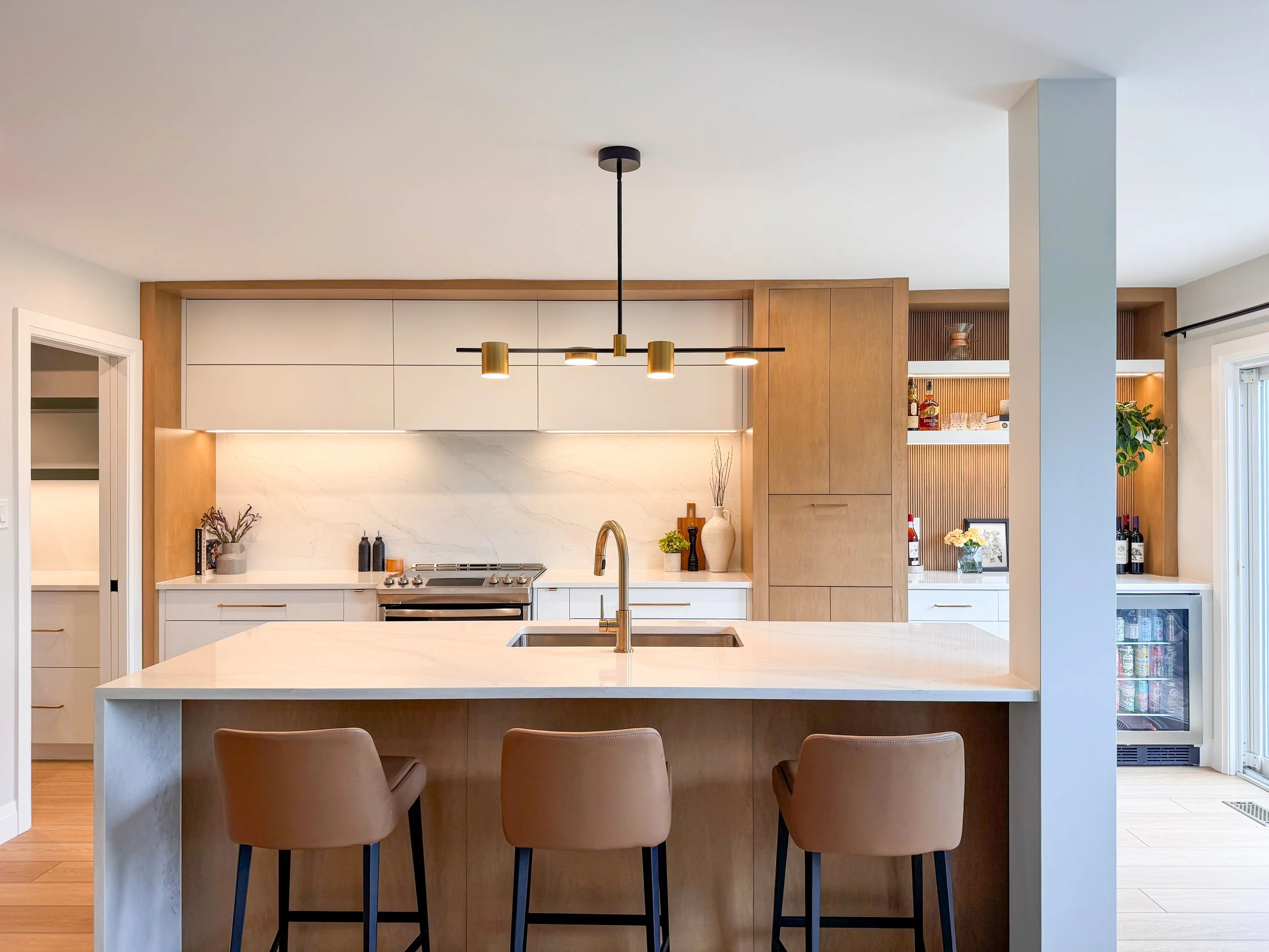 A modern custom kitchen remodel in Waterloo, ON, featuring warm wood cabinets with white slab uppers, a large quartz waterfall island with leather stools, sleek minimal lighting, and a crucial built-in glass-front beverage center for hosting.