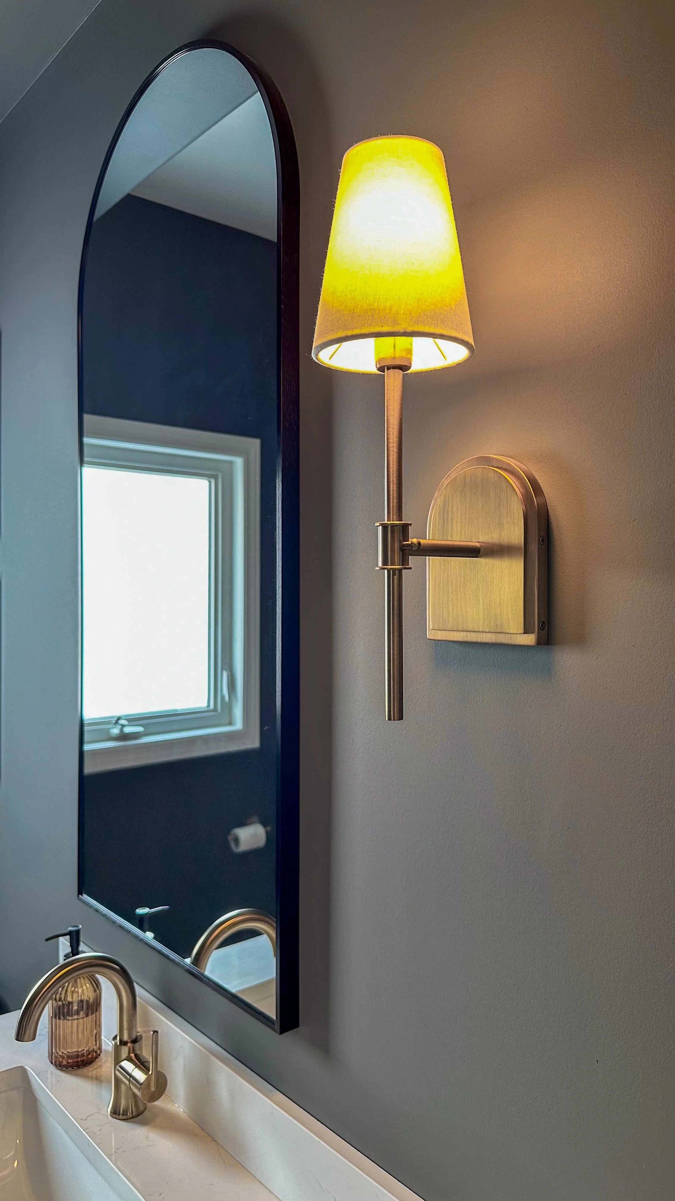 Warm ambient lighting from this brushed brass wall sconce softens the moody dark walls and highlights the elegant curve of the black arched vanity mirror.