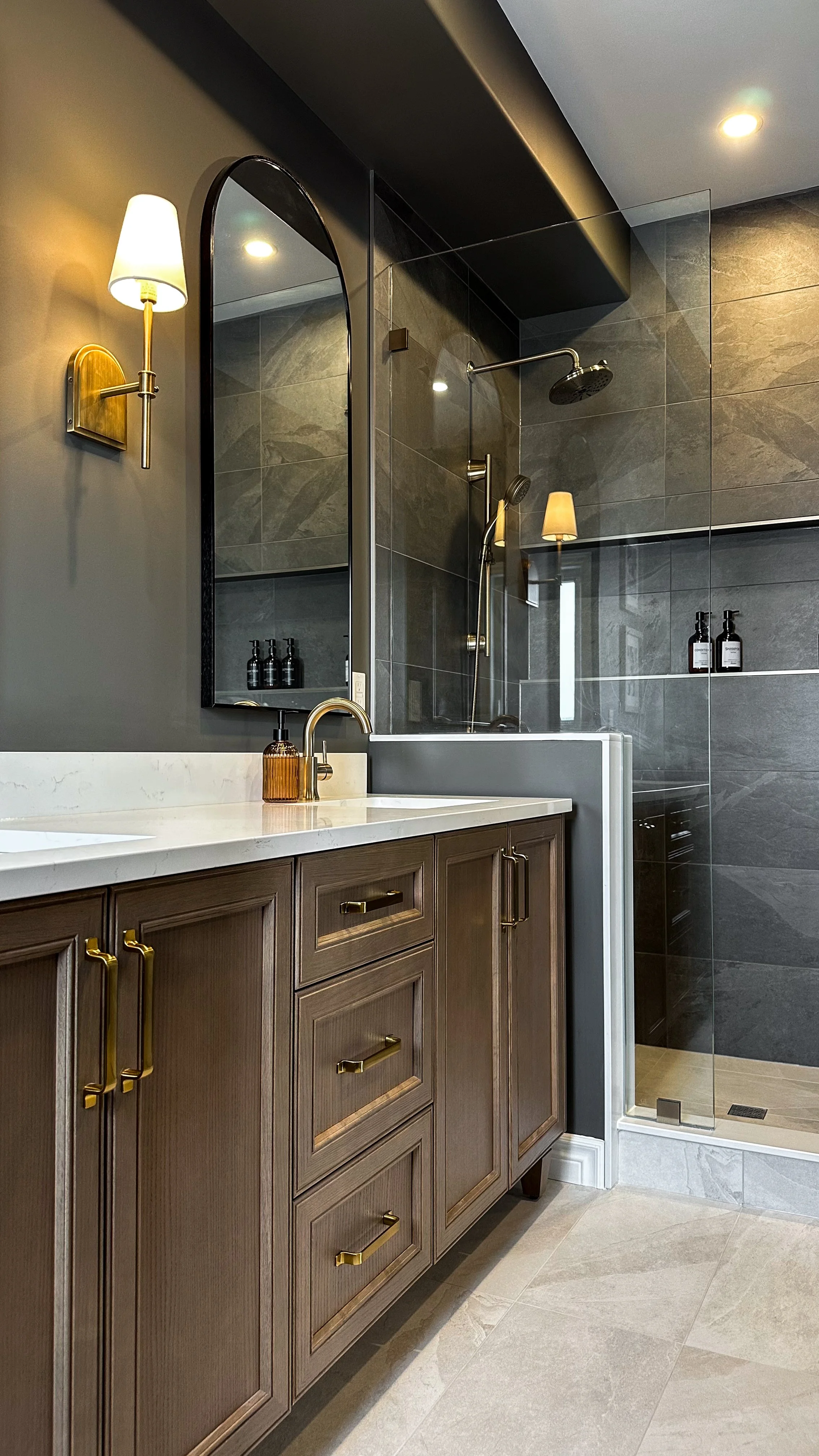 This organic modern double vanity balances warm wood cabinetry and elegant brushed brass hardware against a dramatic dark wall, complete with black arched mirrors and bright white quartz countertops.