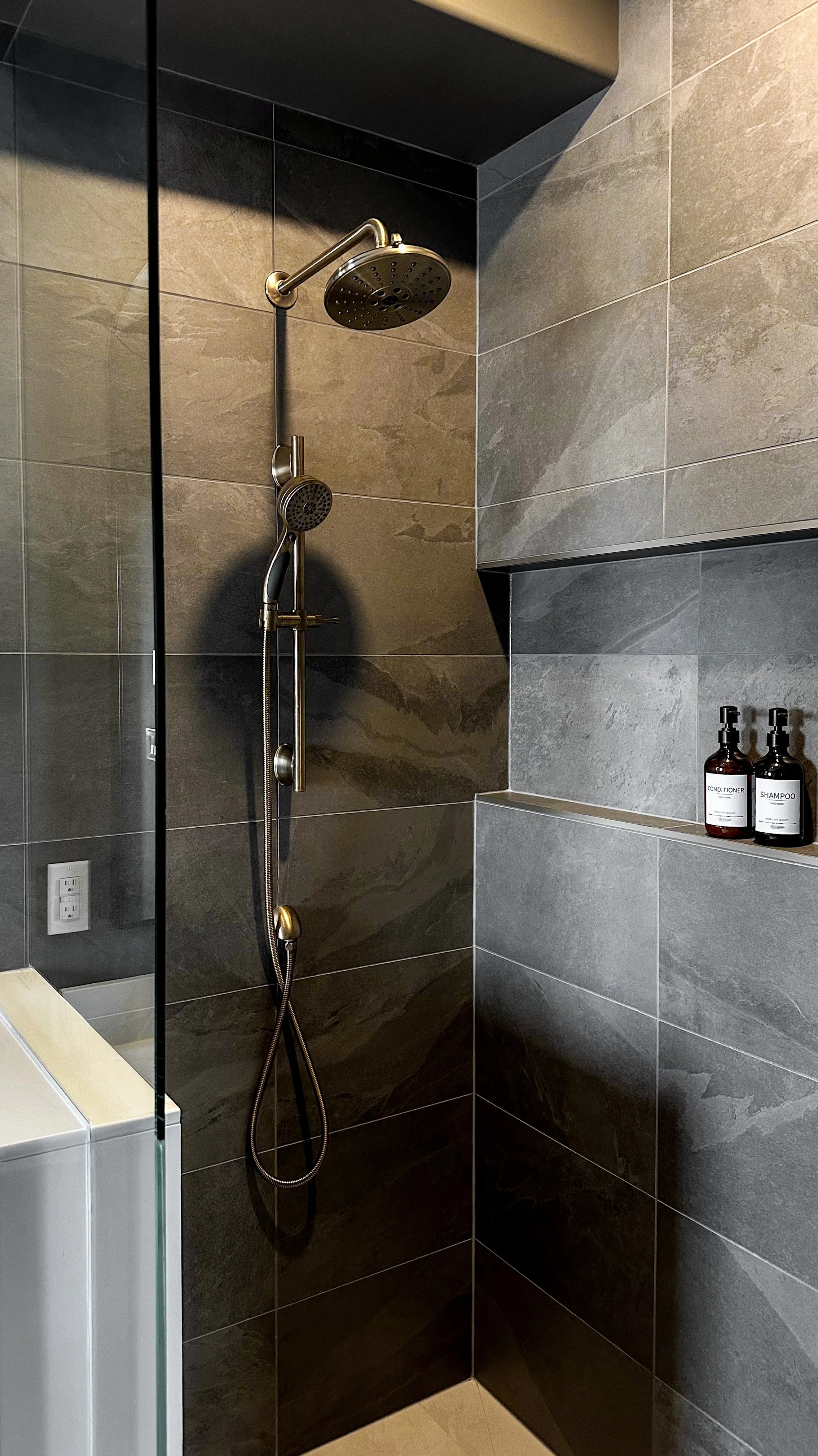 Luxurious brushed brass fixtures, including a rain shower head and handheld sprayer, pop beautifully against the organic texture of the dark grey bathroom shower surround.