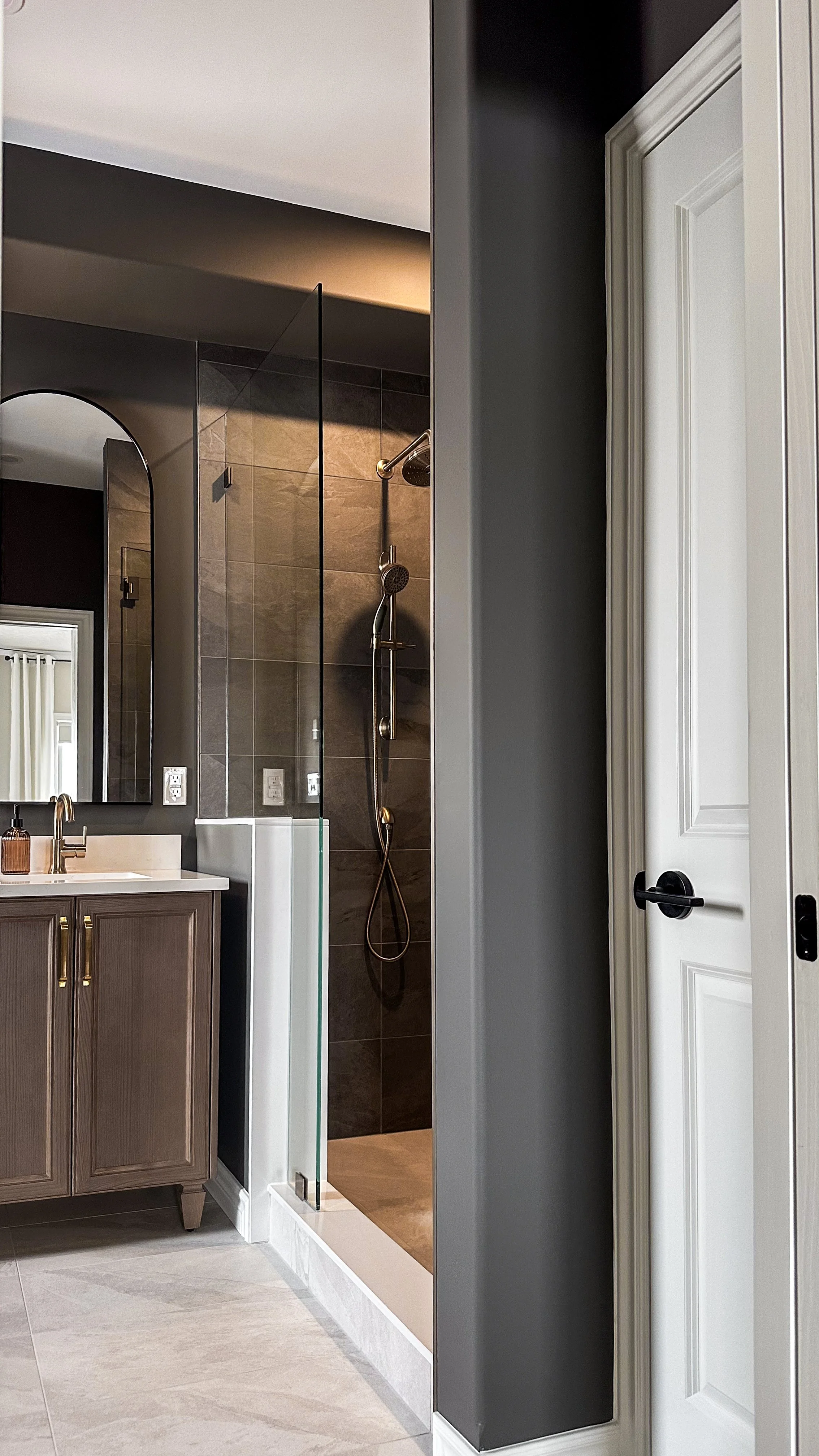 A seamless transition from the bright bedroom into a moody, spa-like ensuite bathroom. The space features rich contrasting tones, a custom wood vanity, and a walk-in glass shower.