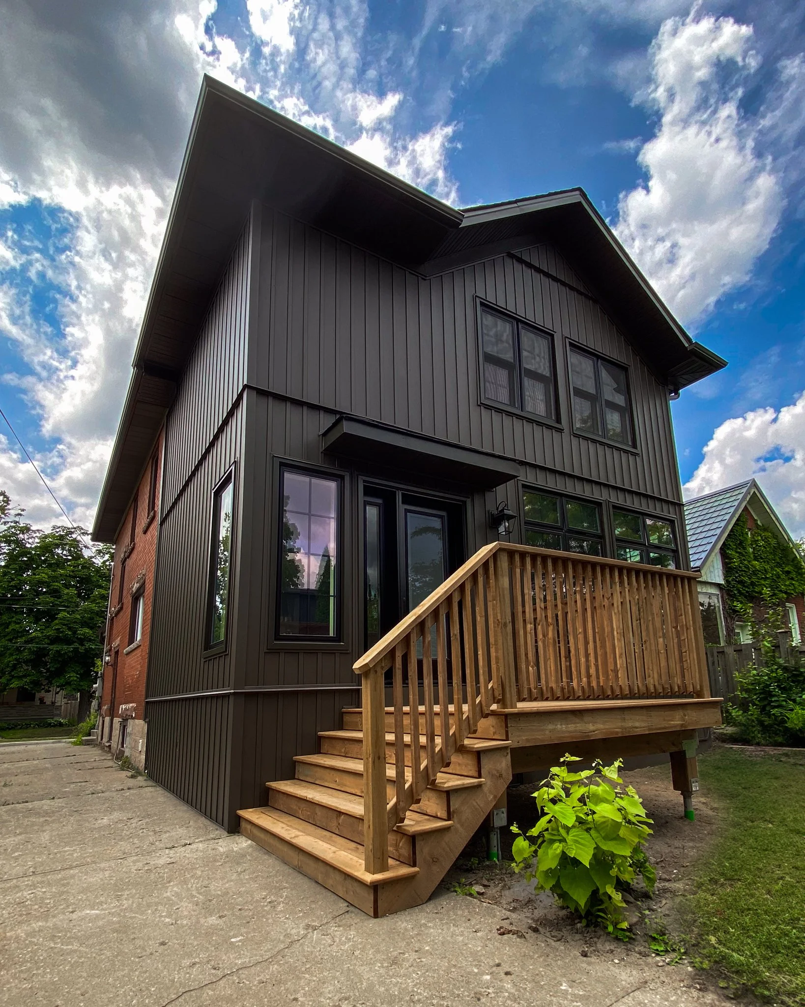 Modern dark-siding two-story home addition with a natural wood deck built onto a traditional red brick house in Kitchener, Ontario.