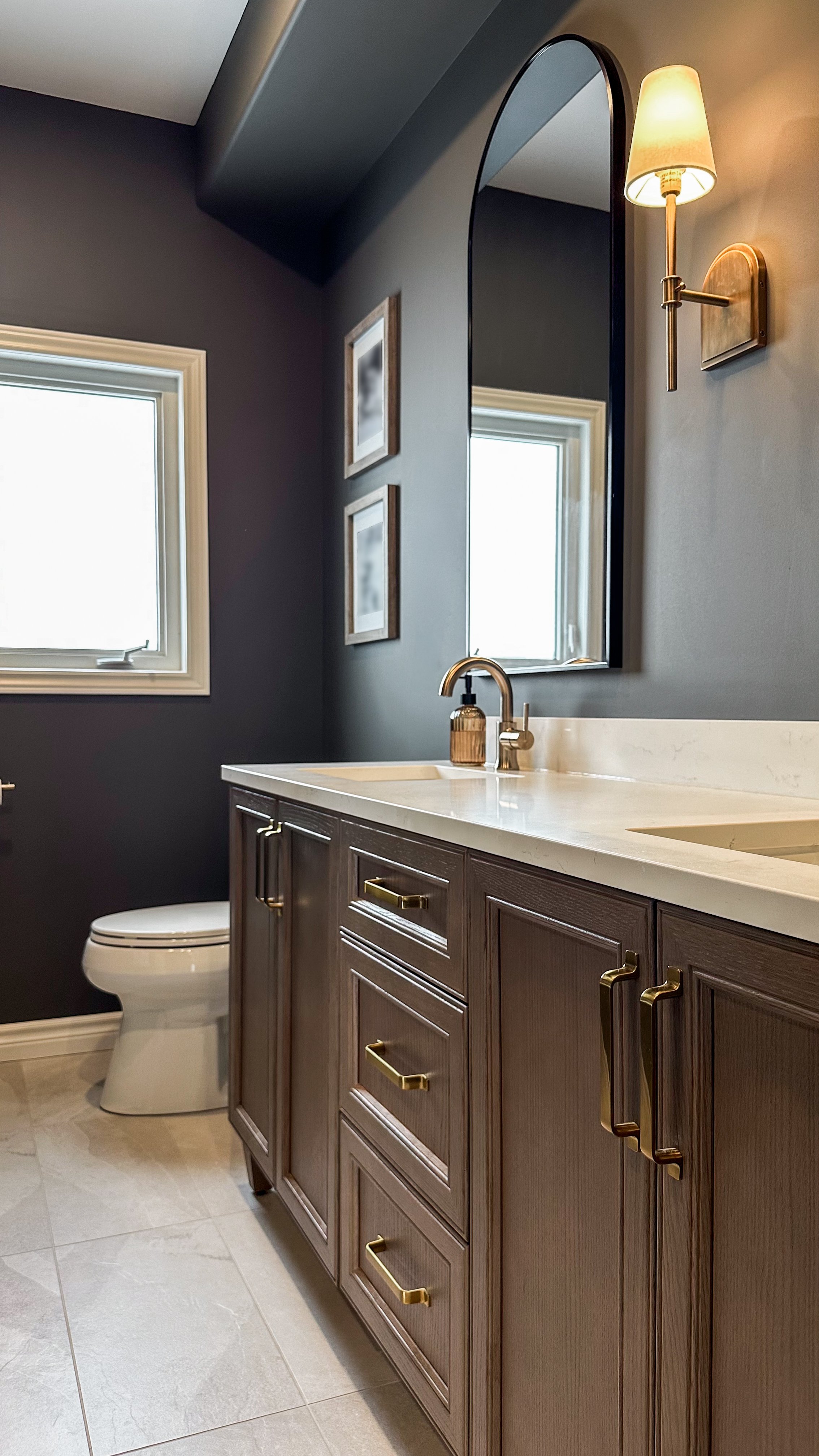 The rich, warm tones of this semi custom wood vanity ground the moody bathroom design, creating a striking contrast with the dark walls, brushed brass drawer pulls, and bright white window trim.