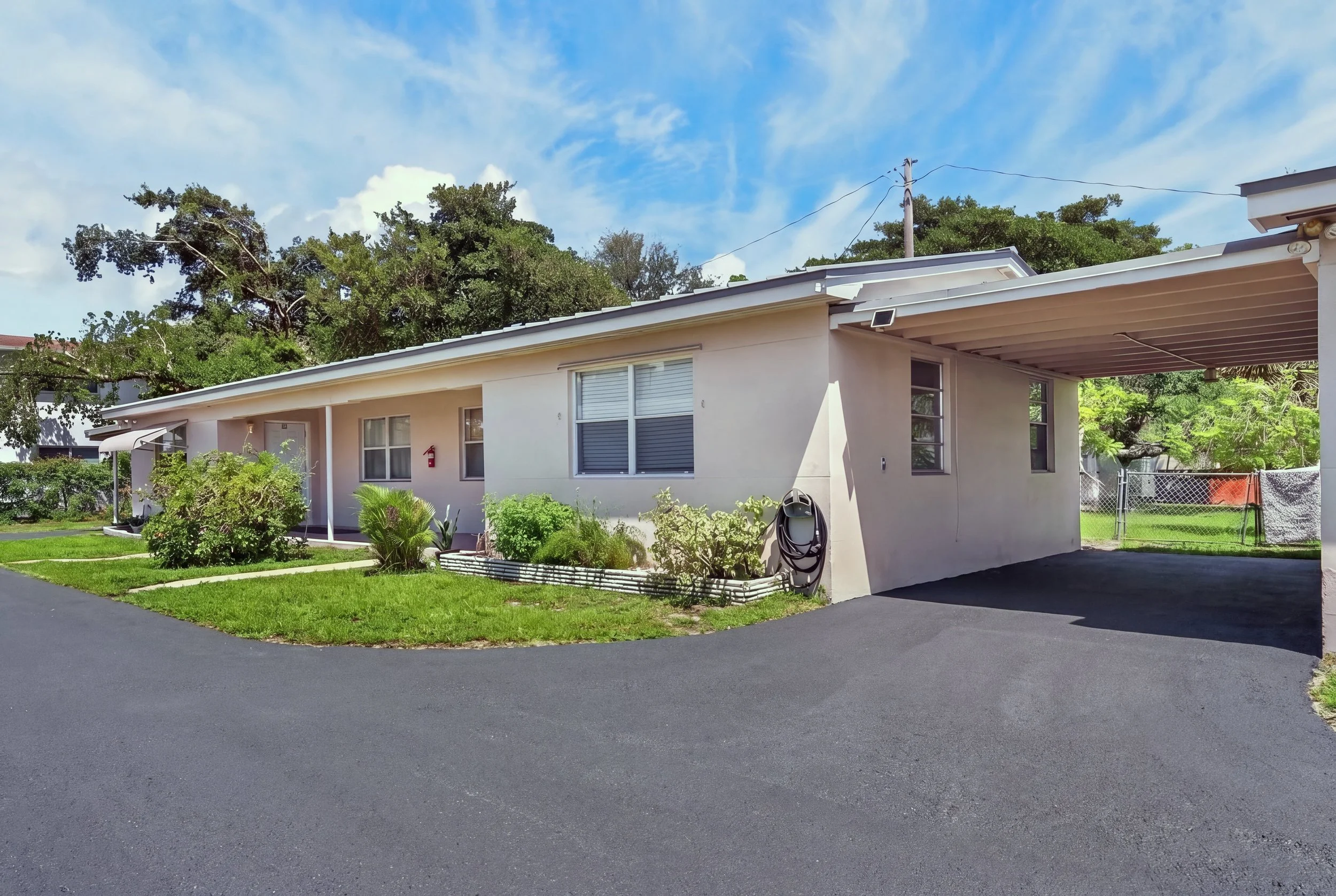 A single-story house with a white exterior, large windows, a covered carport, surrounding greenery, and a bright blue sky with some clouds.