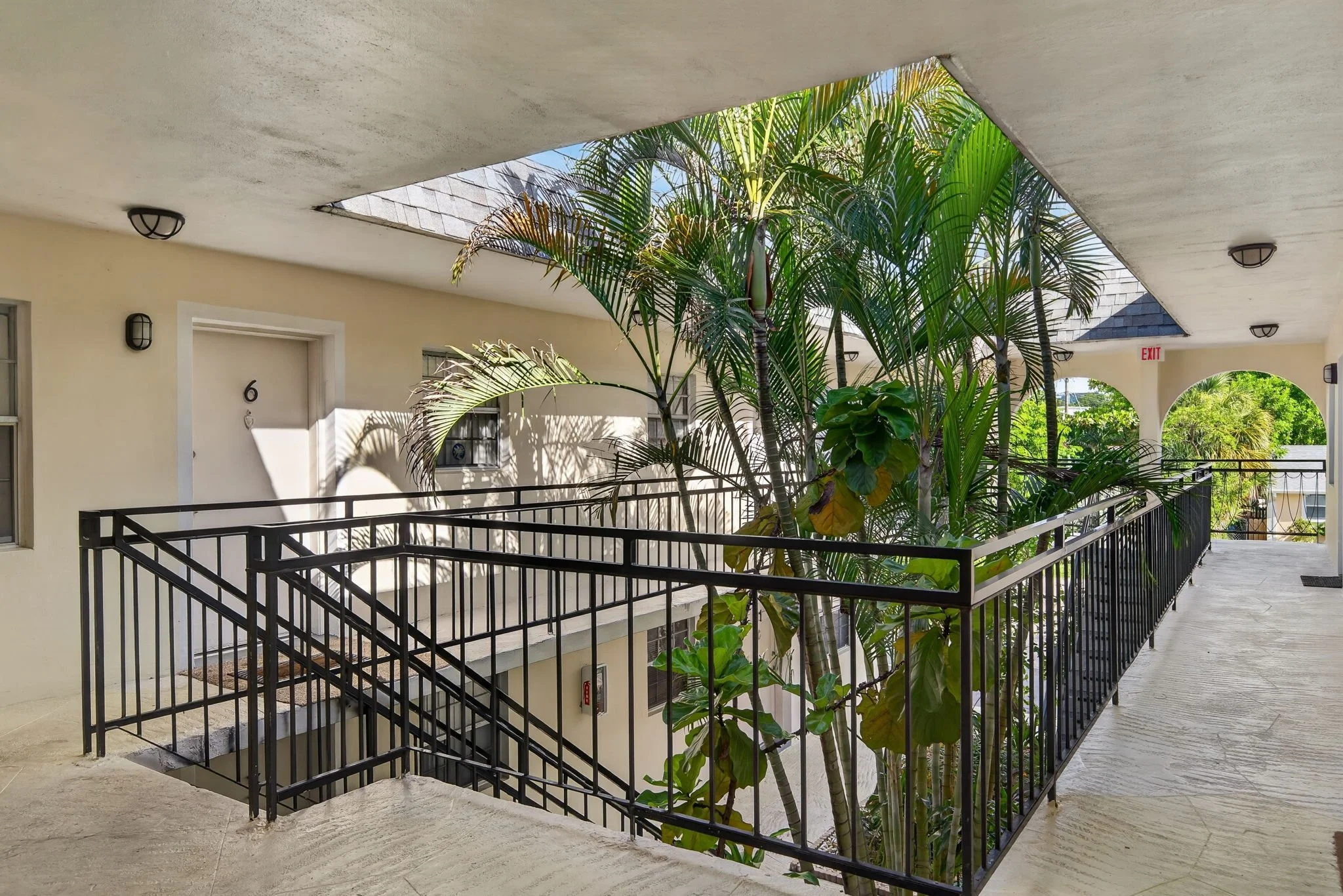 View of a balcony with tropical plants and shadows cast by the trees, with buildings and blue sky in the background.