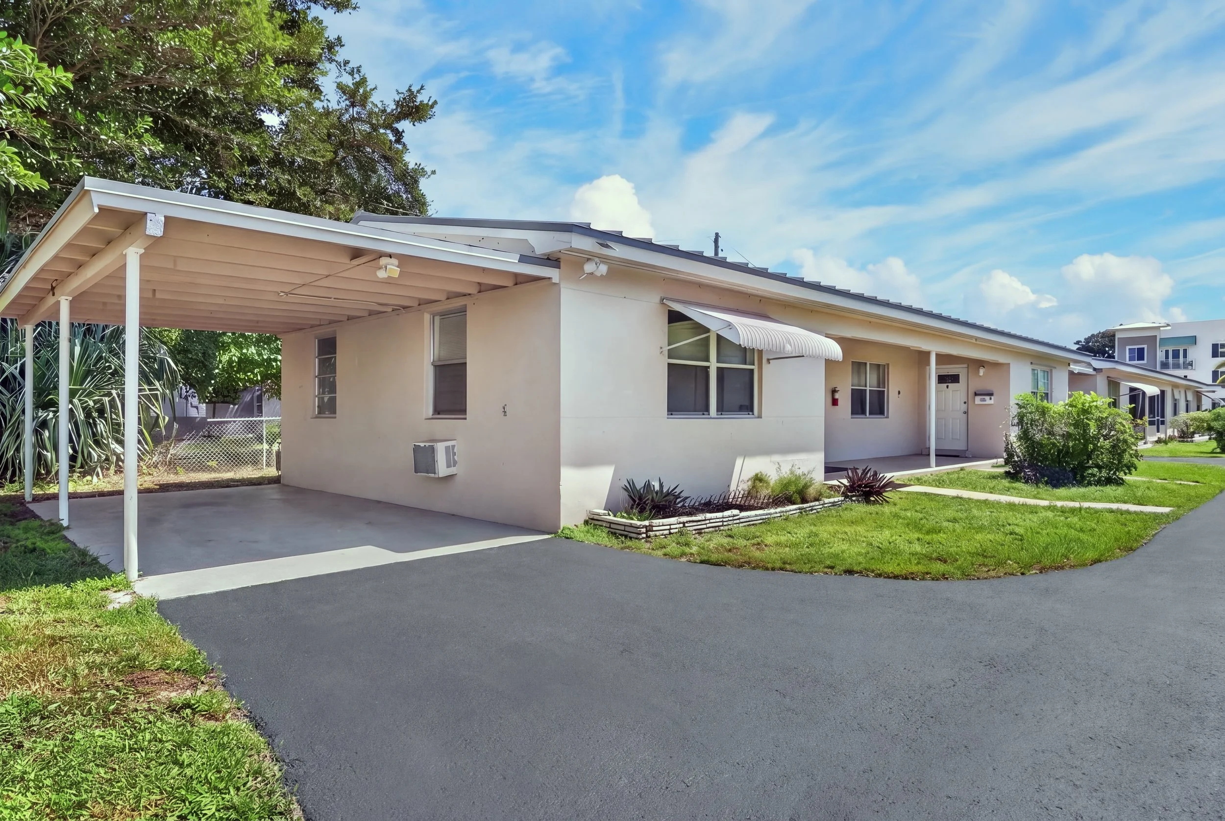 A suburban house with a white exterior, a covered carport, and a well-maintained lawn and garden, under a blue sky with scattered clouds.