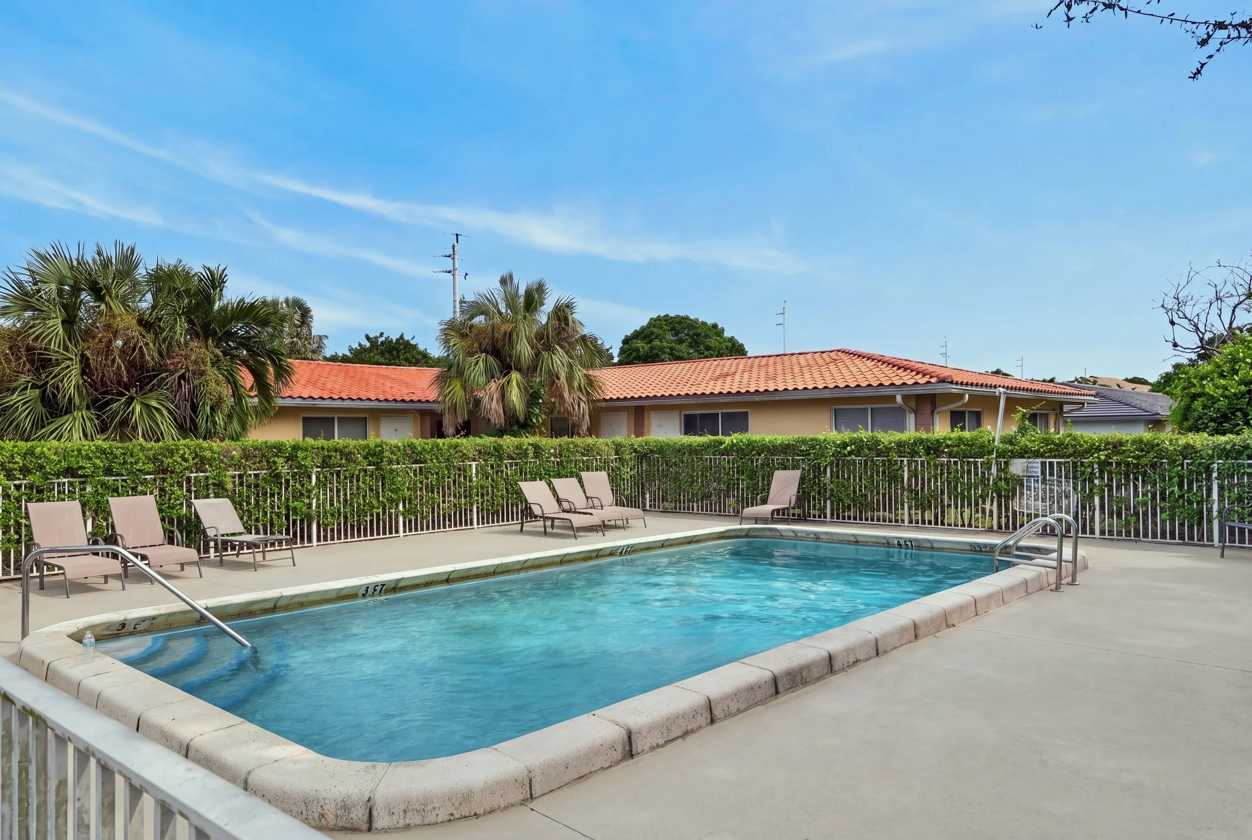 Outdoor swimming pool area with lounge chairs, surrounded by a white fence, with palm and other trees, a building with a red-tiled roof, and a blue sky with some clouds.