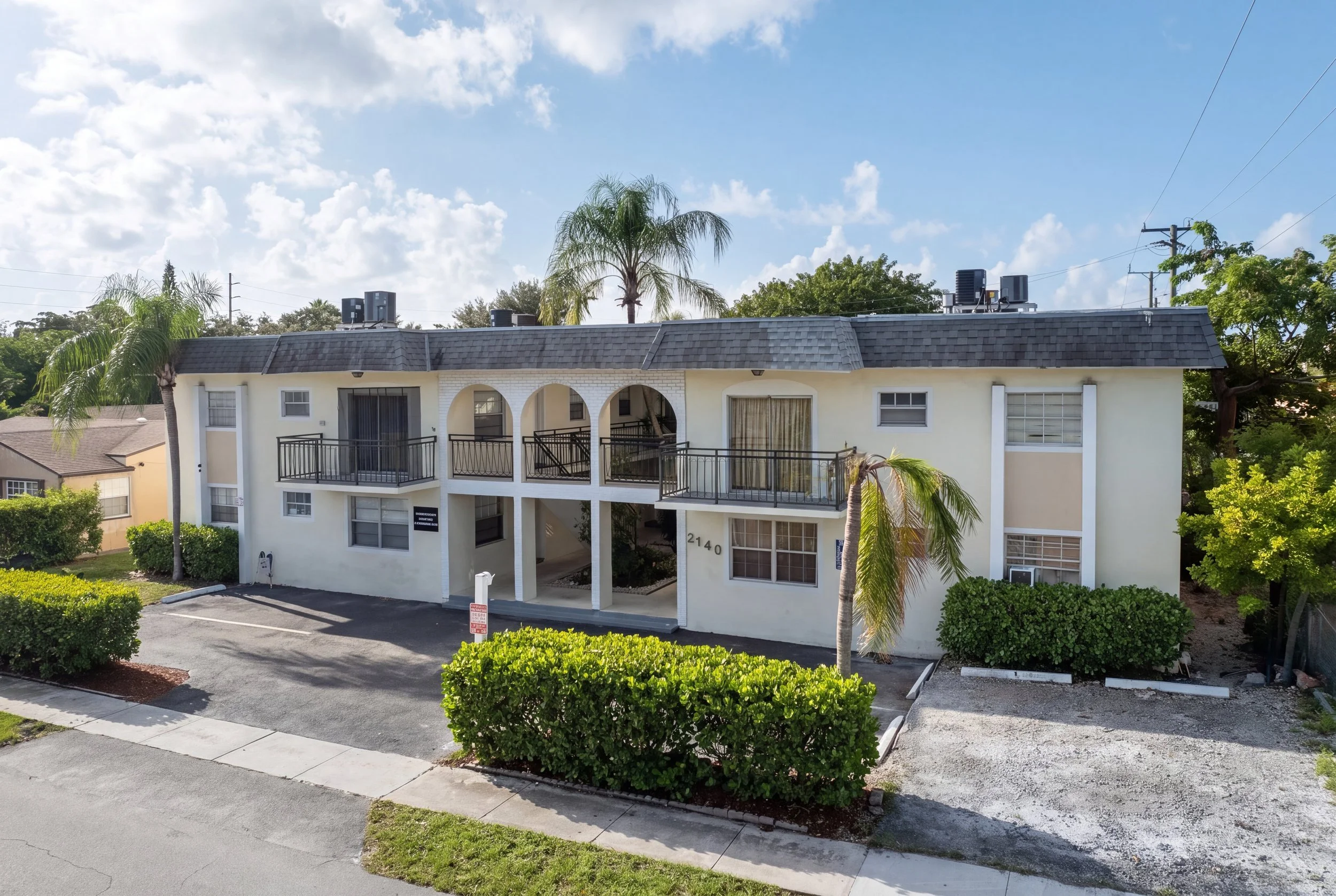 Two-story white residential building with balconies, palm trees, and a parking lot in front, under a partly cloudy sky.