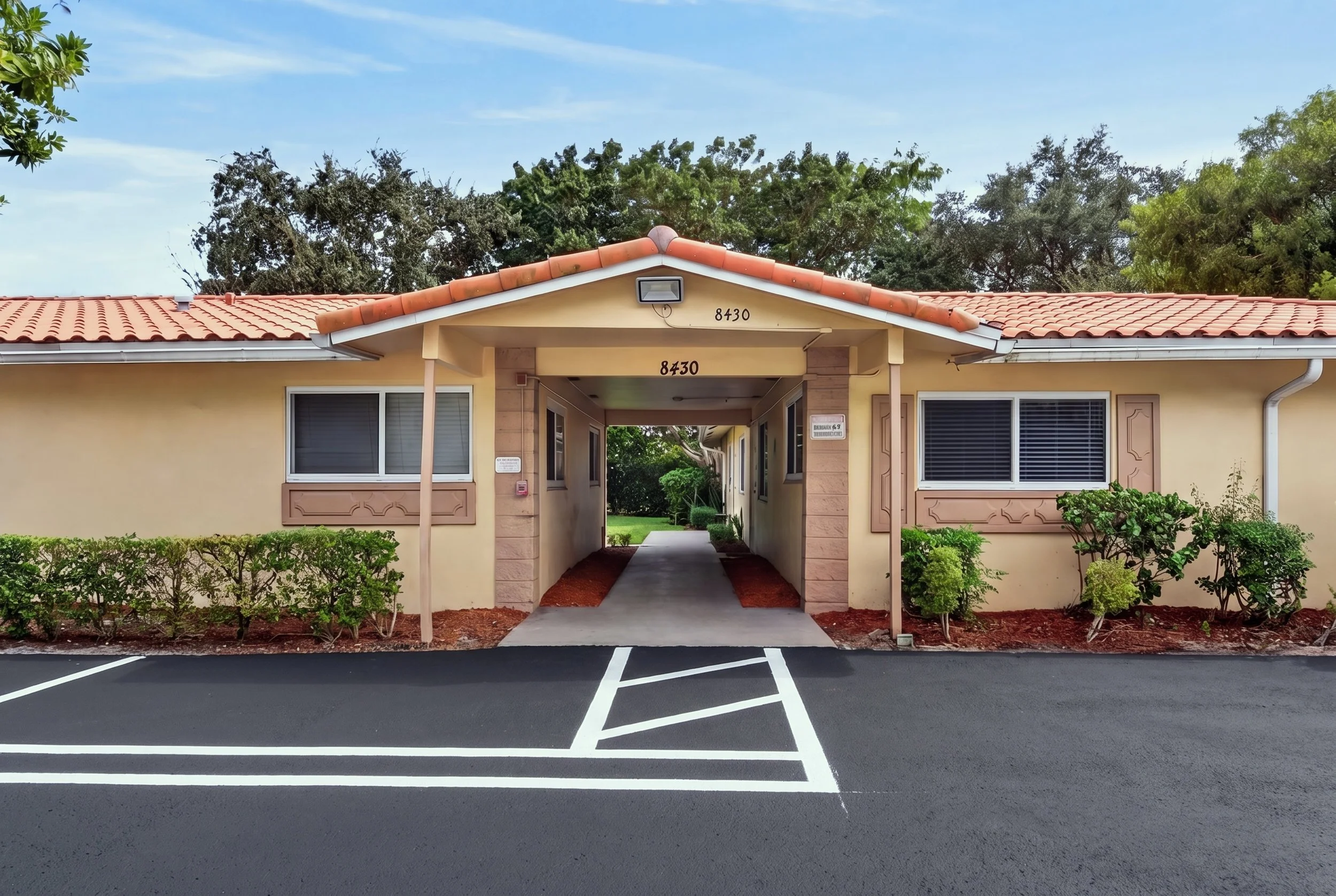 A beige residential building with a tiled red roof and an archway entrance. There are two windows, one on each side of the entrance, with shutters. The building has house numbers 8430. In front, there's a parking lot with white lines and a row of small green bushes along the building. Green trees and a blue sky are in the background.
