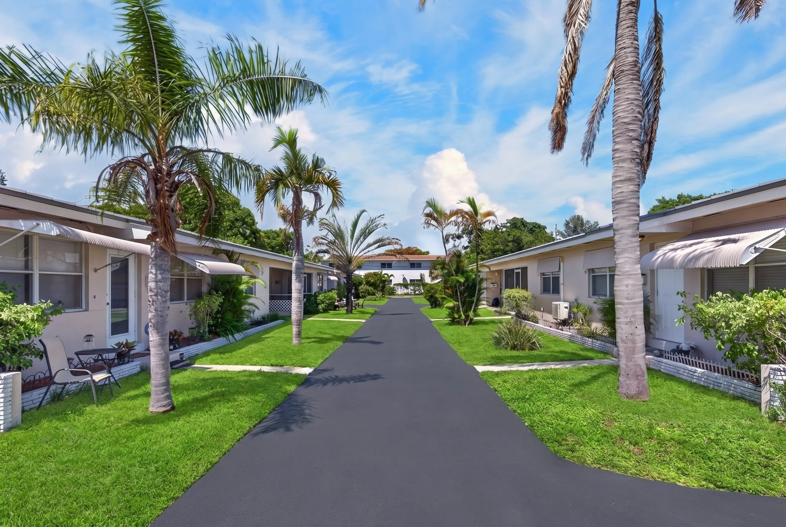A residential courtyard with a paved walkway, surrounded by green lawns, white single-story buildings, and tall palm trees under a partly cloudy sky.