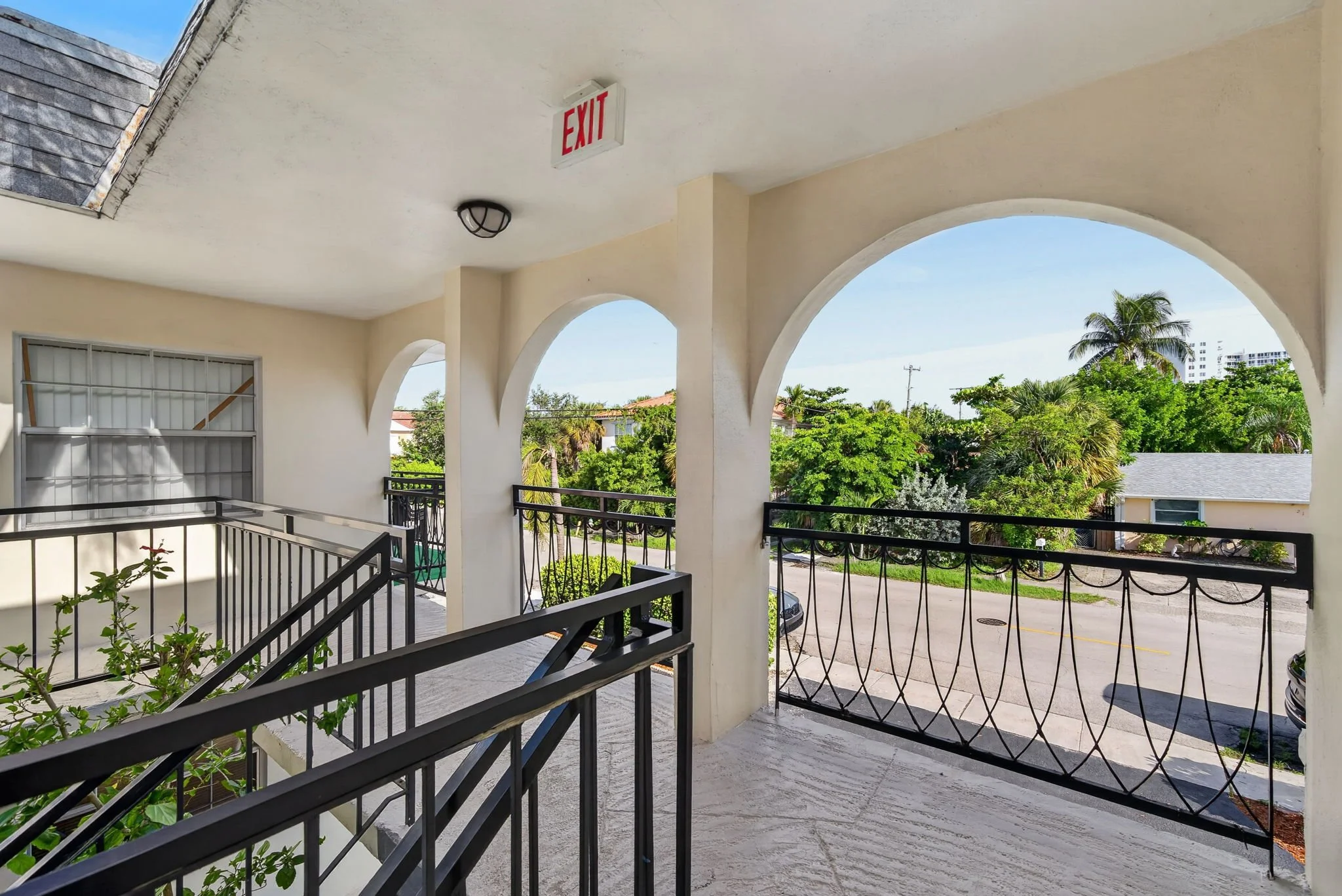 View from a balcony with black metal railings overlooking a street and lush green trees, with a partly cloudy sky and palm trees in the background.