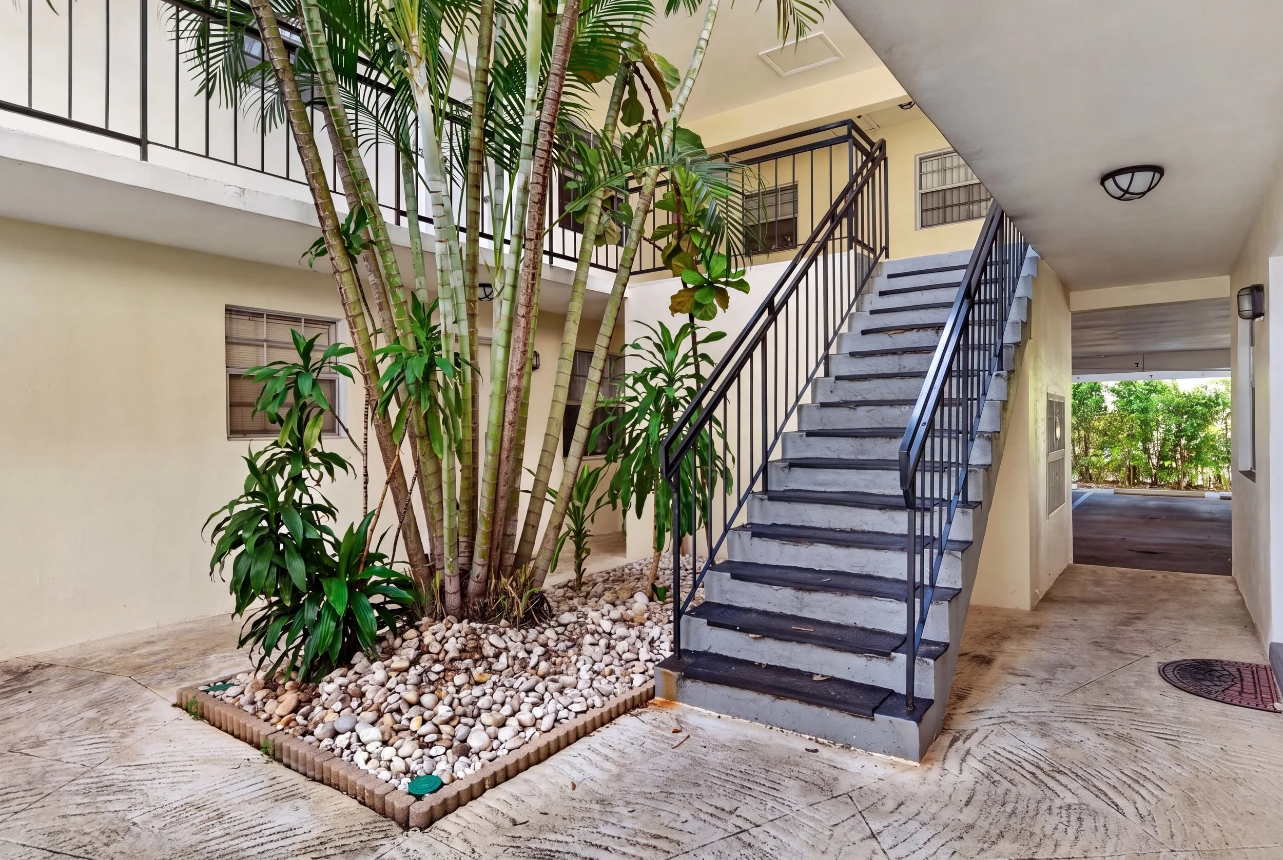 Apartment courtyard with a tall tree, potted plants, and a metal staircase leading to the second floor, with yellow walls and black railing, and window bars visible.