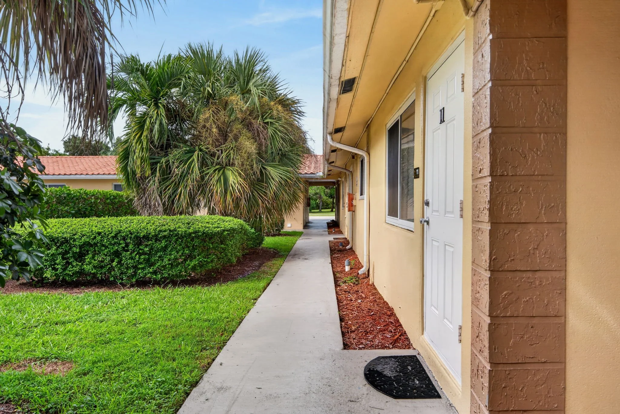 Apartment building entrance with a white door, black mat, concrete pathway, yellow exterior walls, brown brick corner, green bushes, trees, and a blue sky.