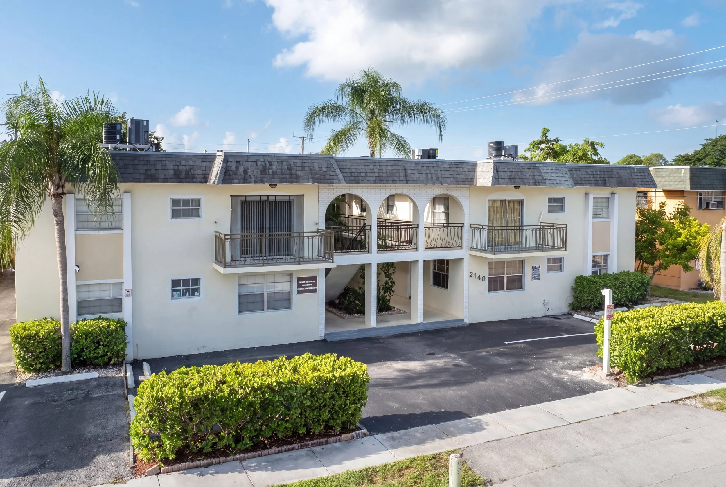 Exterior view of a two-story residential building with white walls, arched balcony openings, and a dark shingle roof, surrounded by green bushes and palm trees, under a blue sky with some clouds.