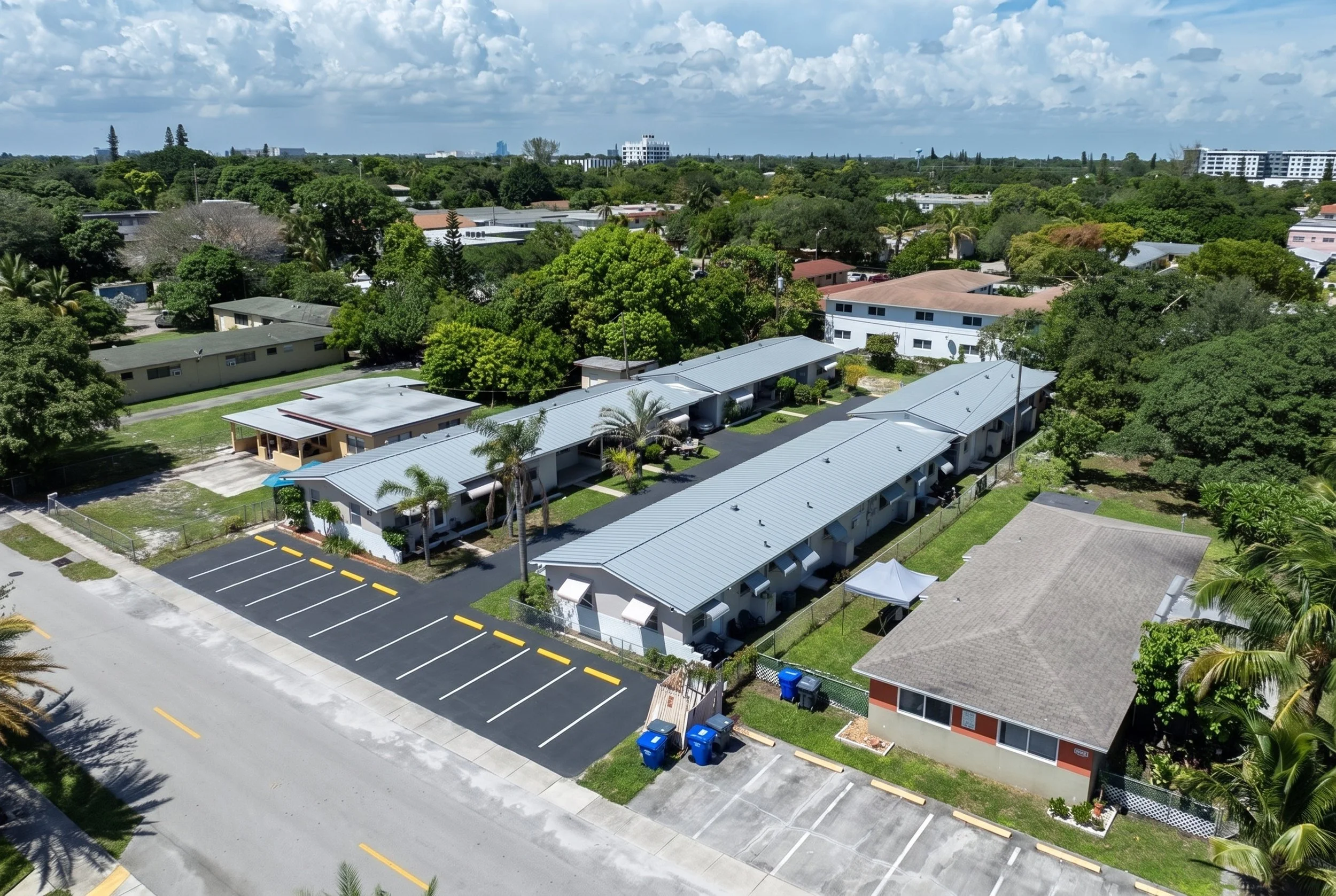 Aerial view of residential buildings and streets with lush green trees under partly cloudy sky.