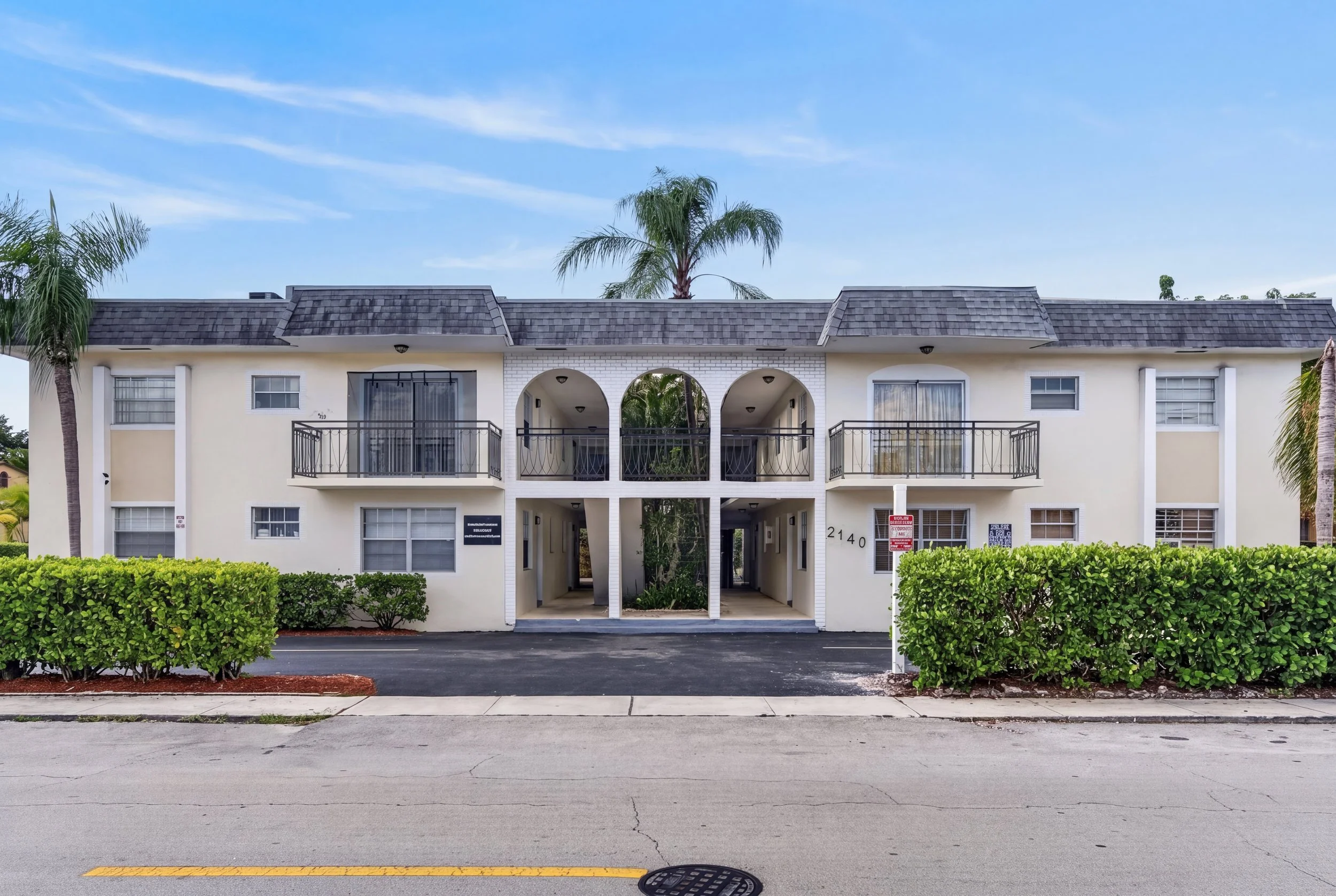 A two-story white apartment building with arched balconies, parking area, and palm trees in front, under a blue sky with some clouds.