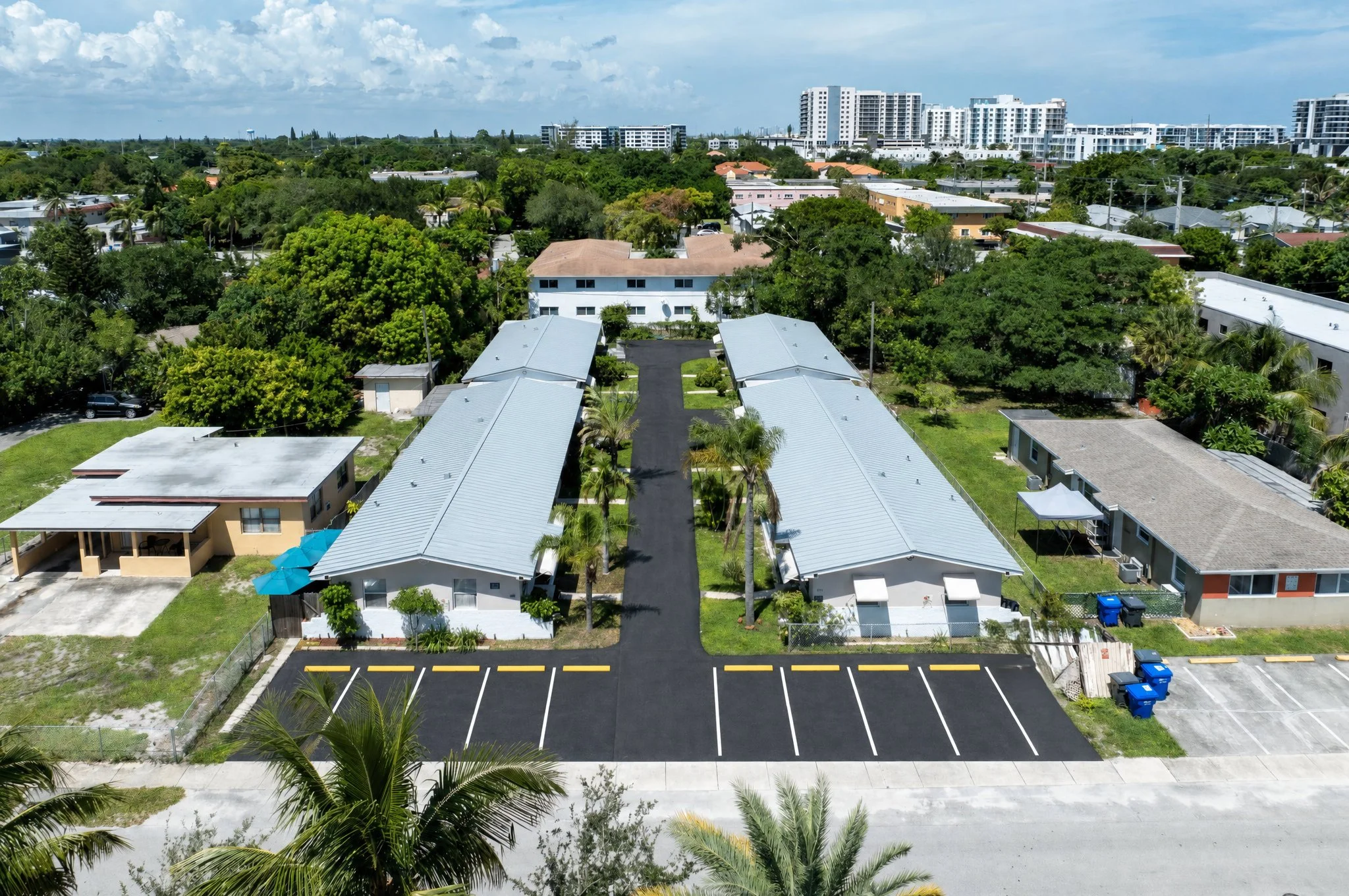 Aerial view of a residential area with single-story houses, parking lots, and tall trees, with modern high-rise buildings in the background under a partly cloudy sky.