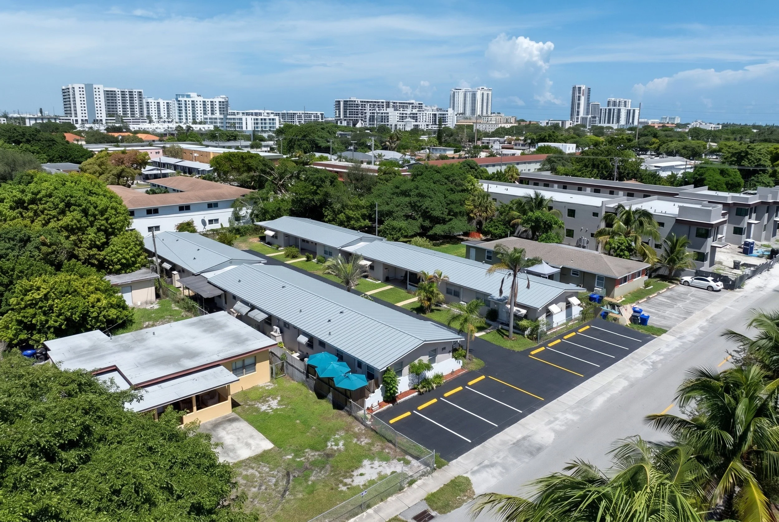 Aerial view of a residential area with single-story houses, surrounded by greenery and trees, with taller apartment buildings and a city skyline in the background under a partly cloudy sky.