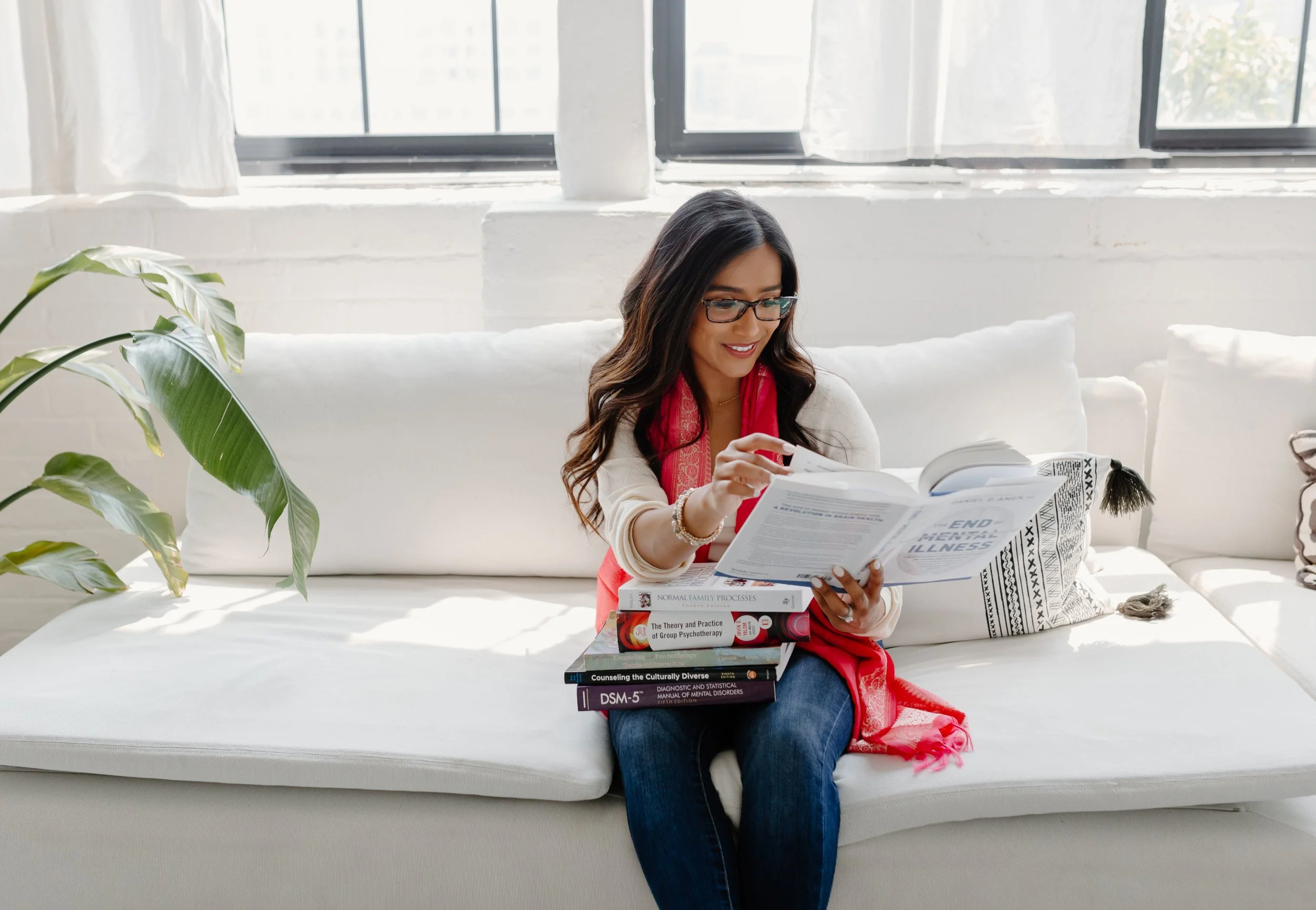 Woman with long dark hair, glasses, and a red scarf reading a book on a white sofa with a stack of psychology and therapy books nearby in a bright, airy room.