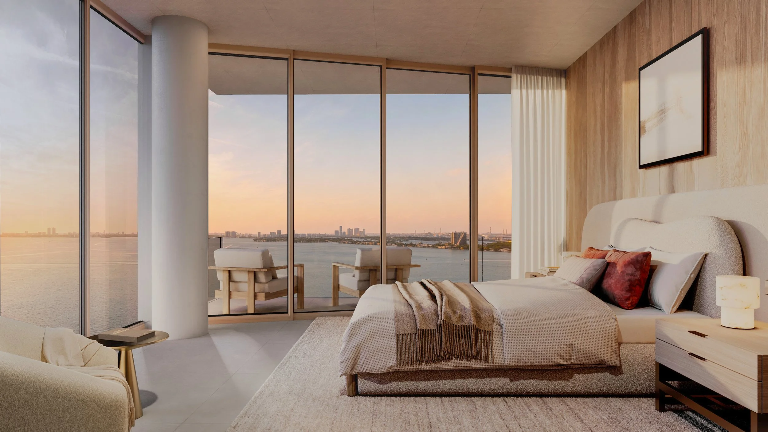 Modern bedroom with floor-to-ceiling windows offering a view of a river and city skyline at sunset. The room has a large bed with beige bedding and decorative pillows, a bedside table with a lamp, and seating area on the balcony with chairs.