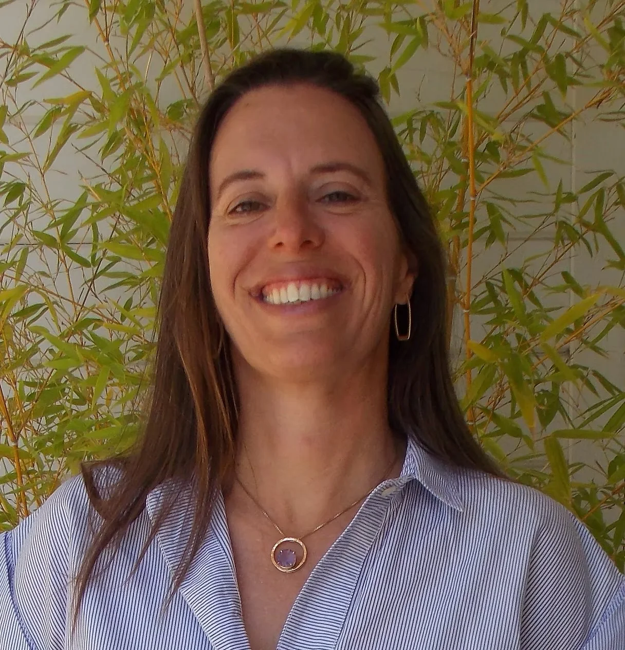 A woman with long brown hair smiling, wearing a striped shirt, jewelry, and earrings, standing outdoors in front of a leafy green plant. Downtown Novato Board Member.