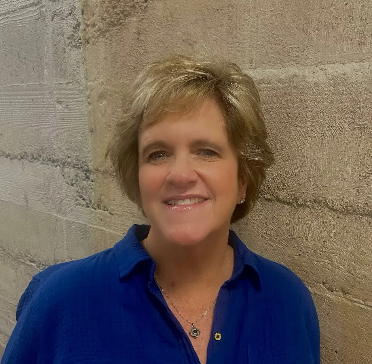 A woman with short, layered blonde hair smiling, wearing a blue collared shirt and a necklace, standing against a beige textured brick wall. Downtown Novato Board Member.