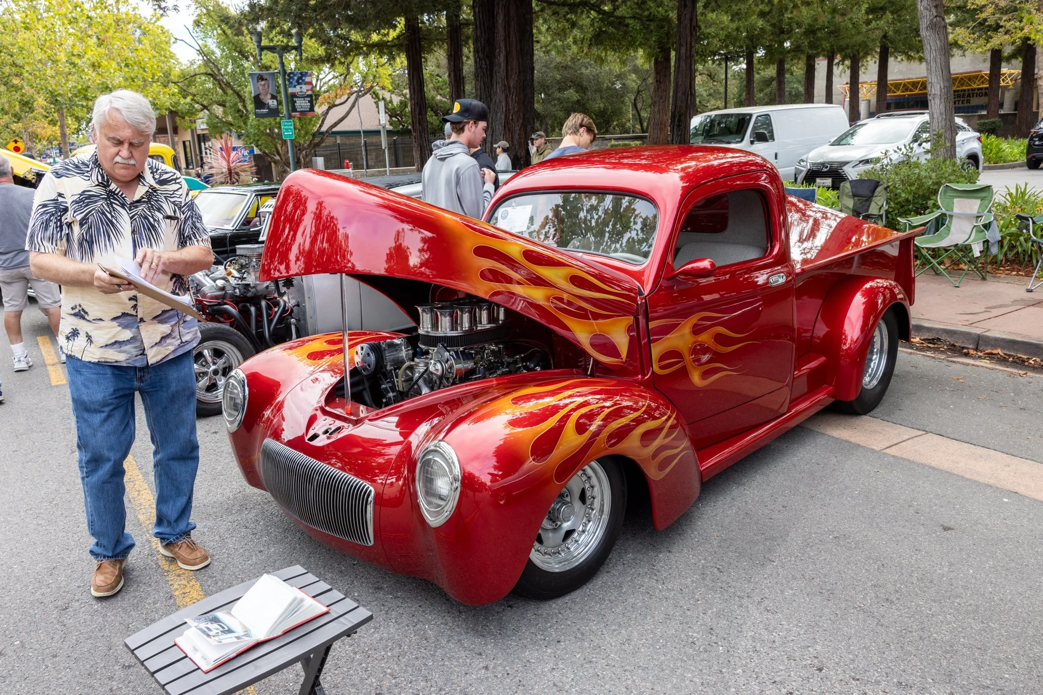 A crowd of people at a car show, with a bright red vintage pickup truck with flames painted on its sides on display, in Downtown Novato California.
